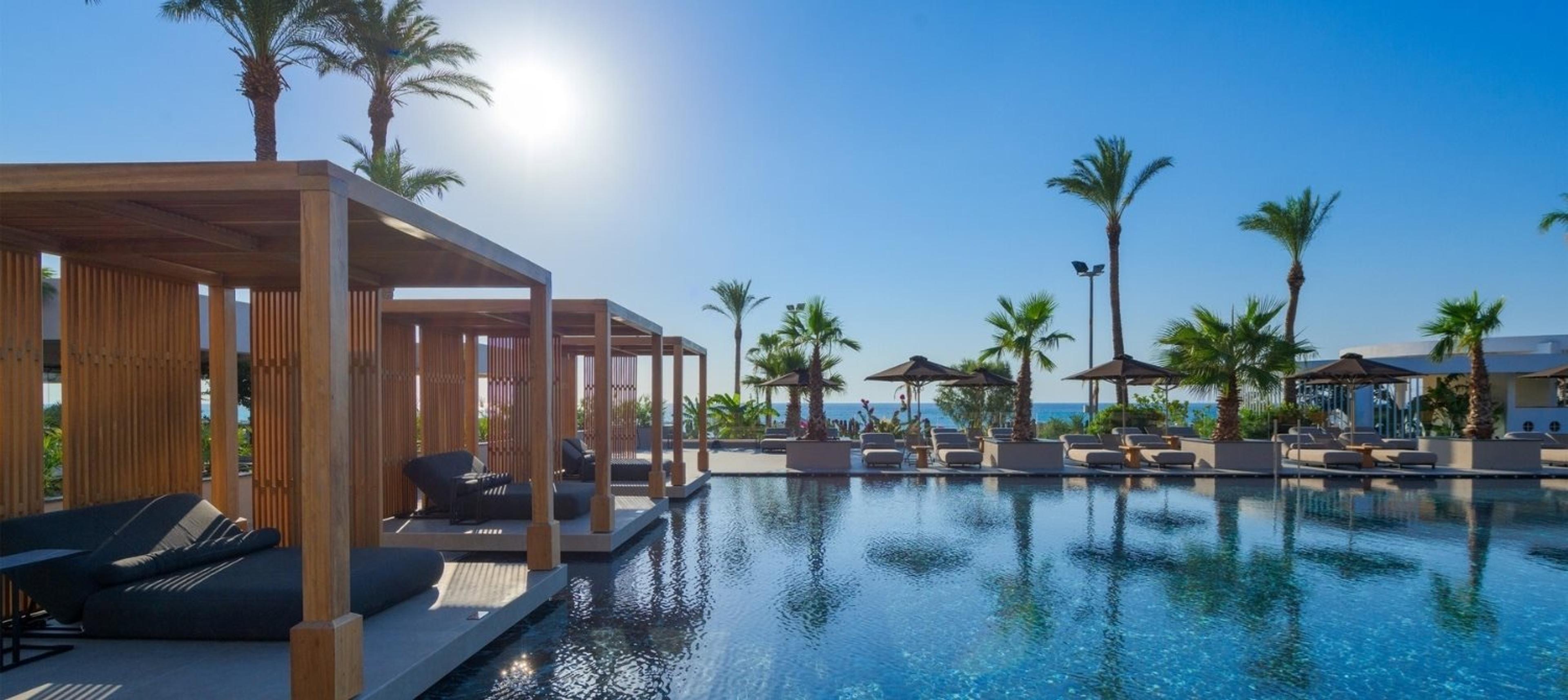 Morning sun flares over an infinity pool edged by wooden cabanas and palm trees, with hills and the sea visible past rows of loungers.