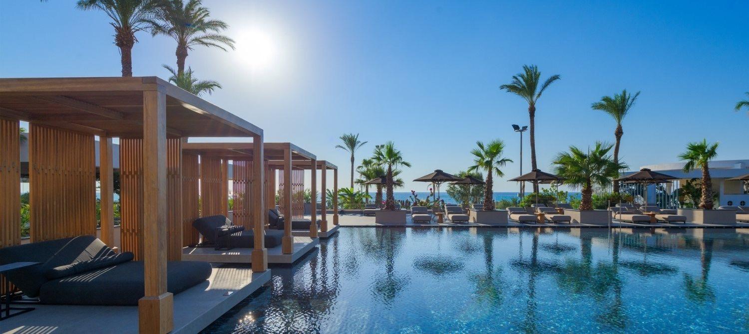 Morning sun flares over an infinity pool edged by wooden cabanas and palm trees, with hills and the sea visible past rows of loungers.