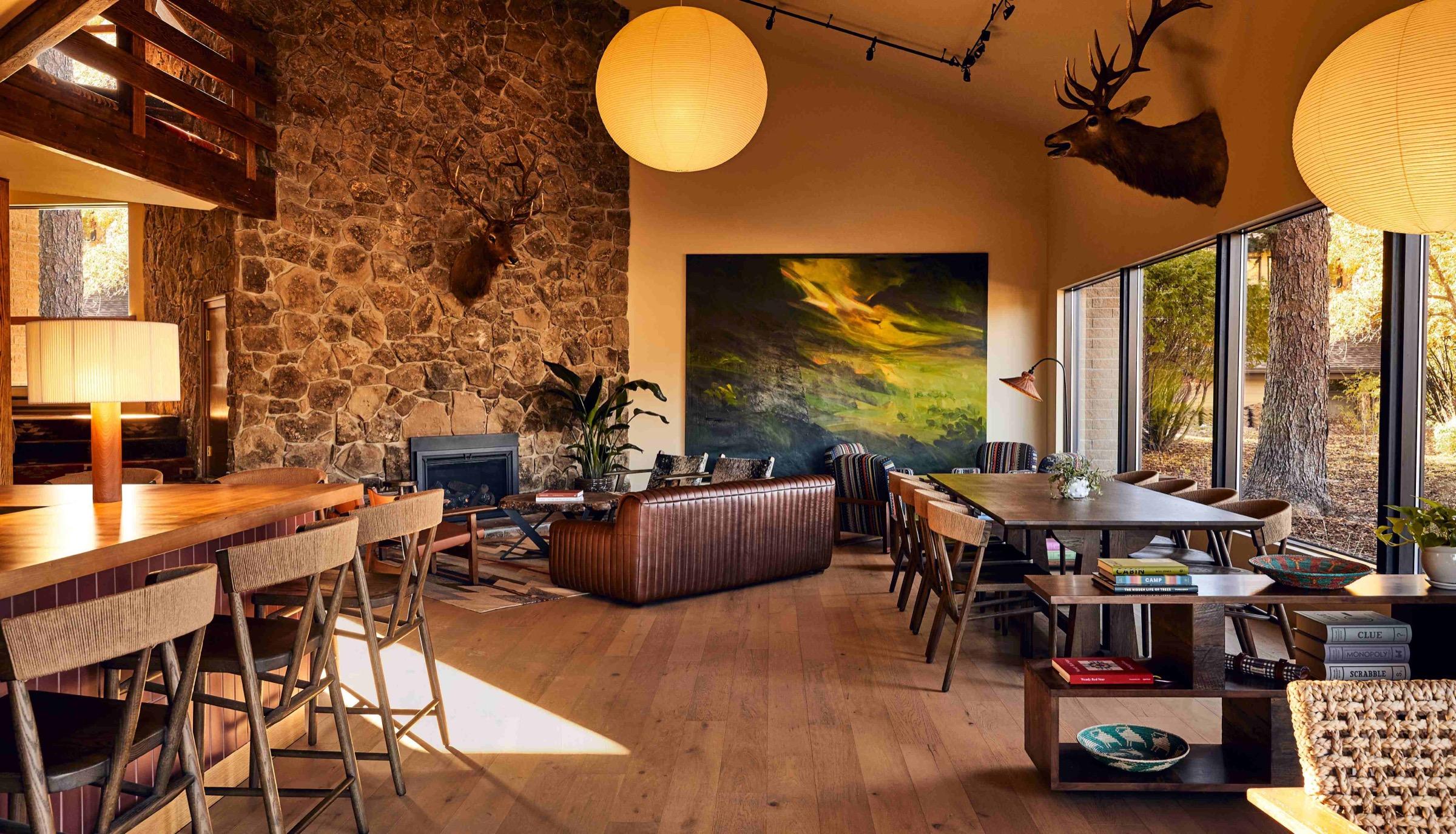 A lodge lounge with a stone fireplace wall, a mounted deer head, a tufted leather sofa and a long communal table, with tall windows onto trees.