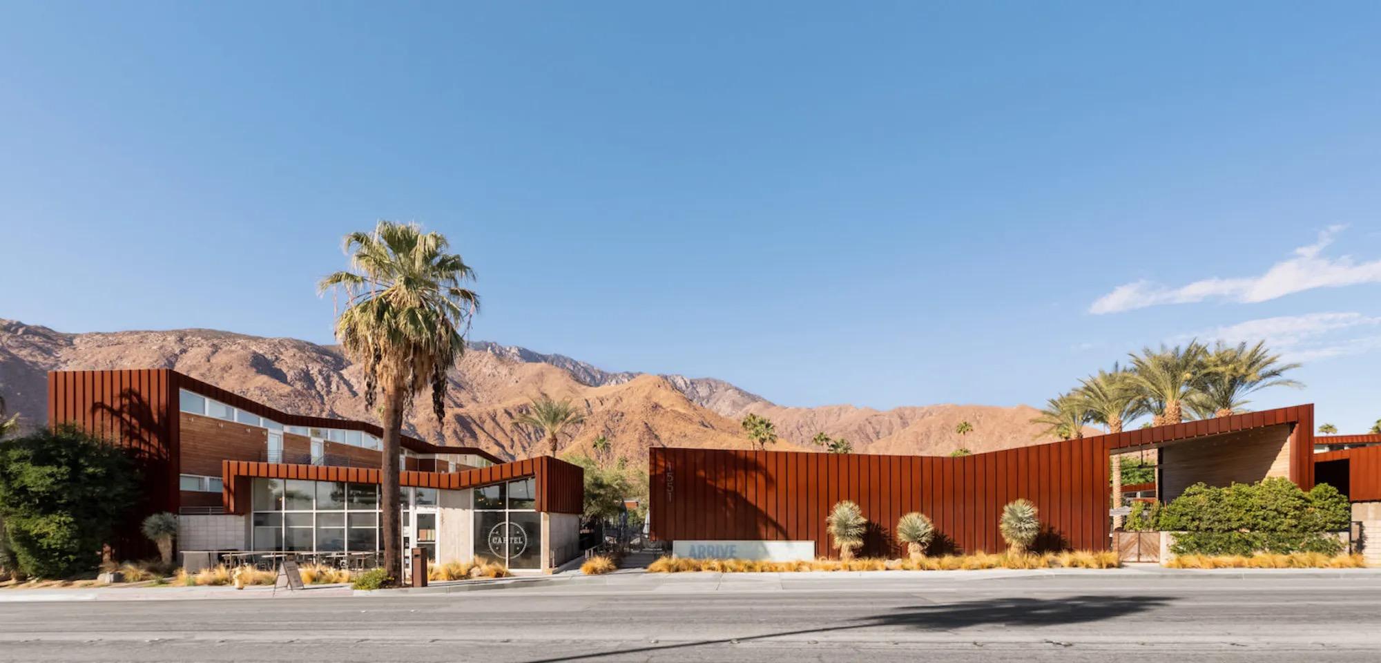The low, rust-coloured front of the hotel under a clear blue sky, with a tall palm on the left, bare brown mountains behind and a row of ornamental grasses along the pavement.