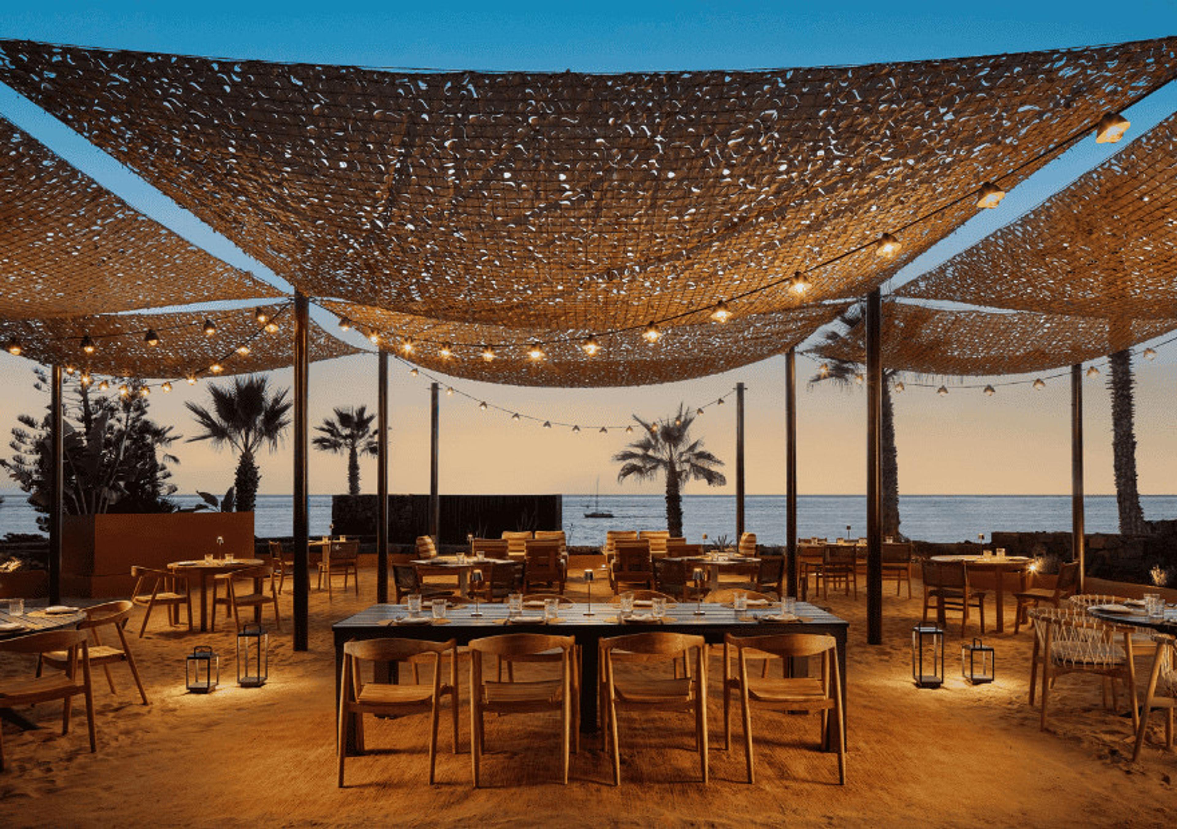 Tables on the sand under woven shades and strung lights at SEEN, the sea beyond at dusk