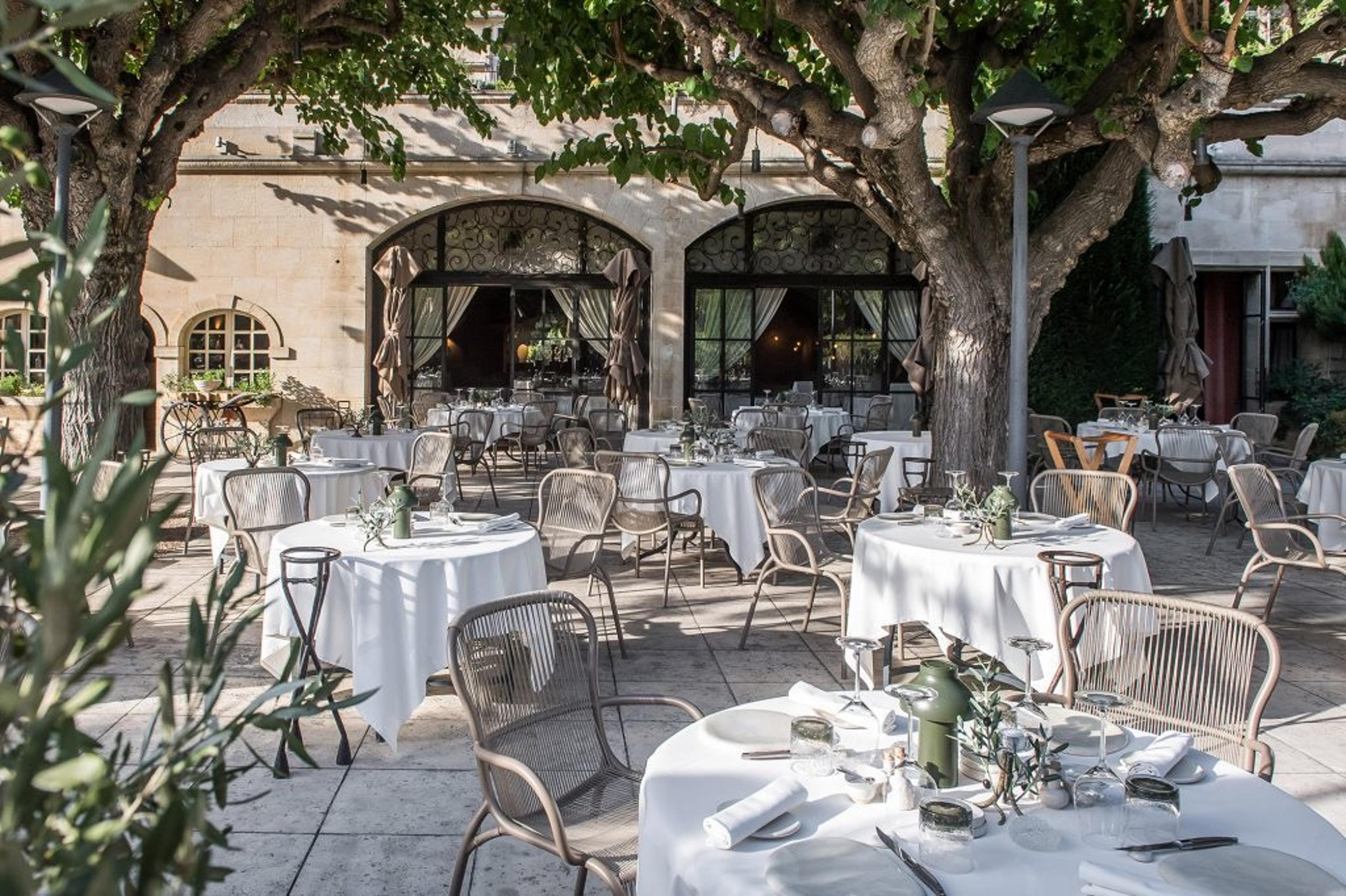 Tables laid in white on a terrace under plane trees, in front of the stone farmhouse.