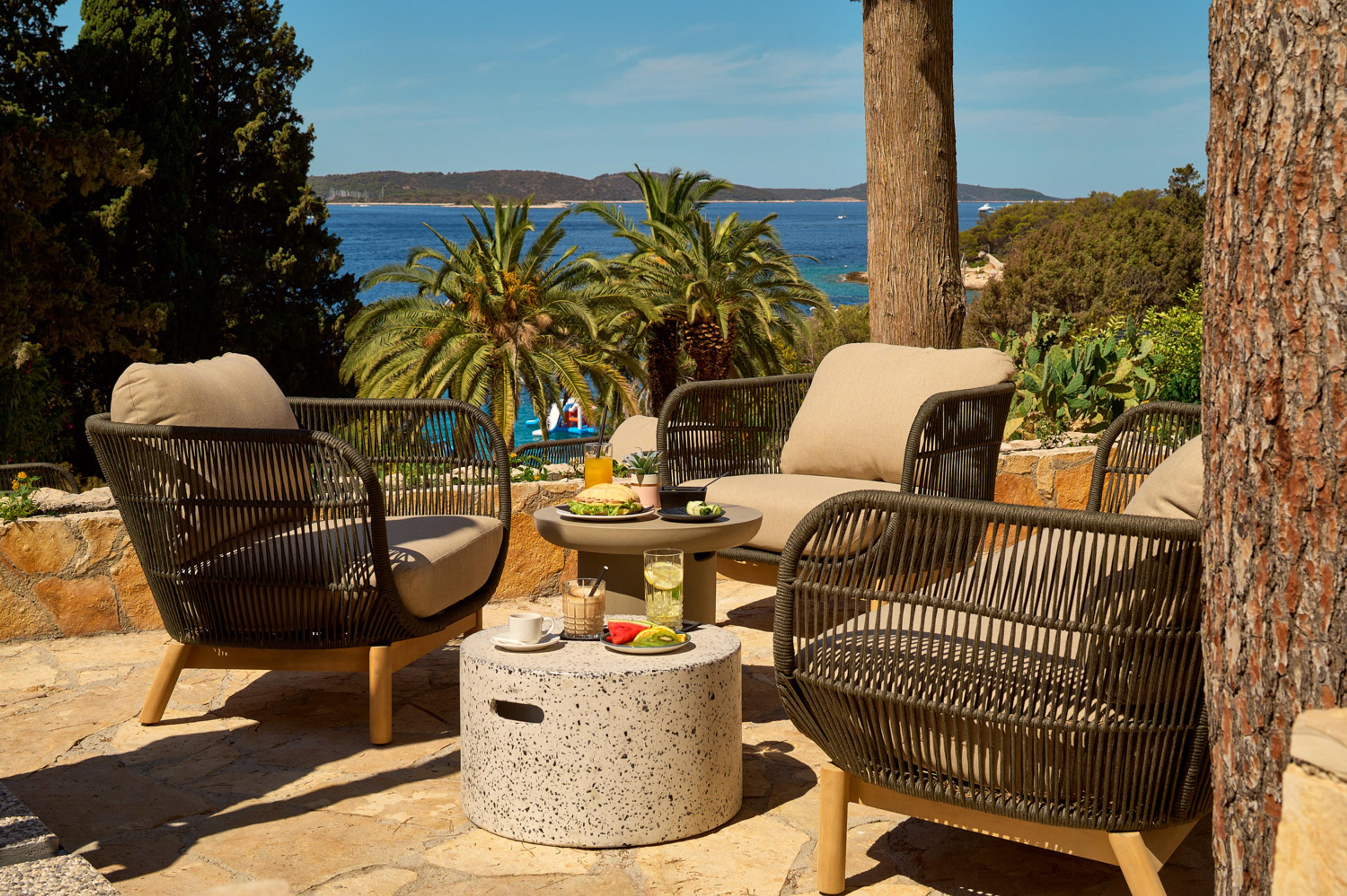 Outdoor lounge chairs around two round tables set with a sandwich, fruit, coffee and drinks, with palm trees and the sea behind.