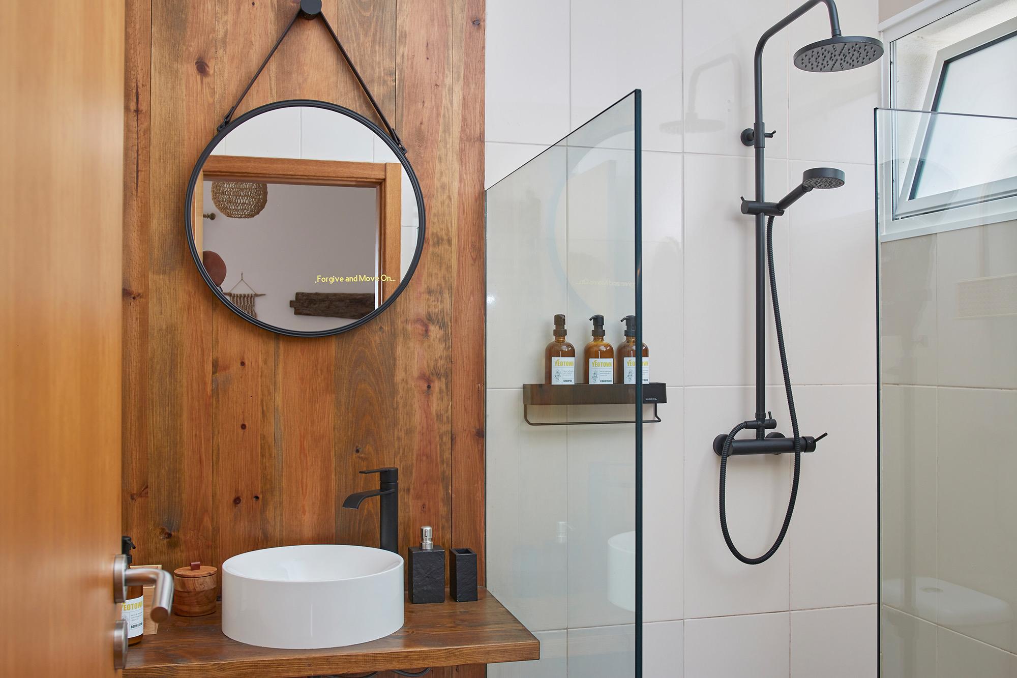 A bathroom with a round mirror on a wood-panelled wall, a vessel sink on a wood counter, and a glass-walled walk-in shower.