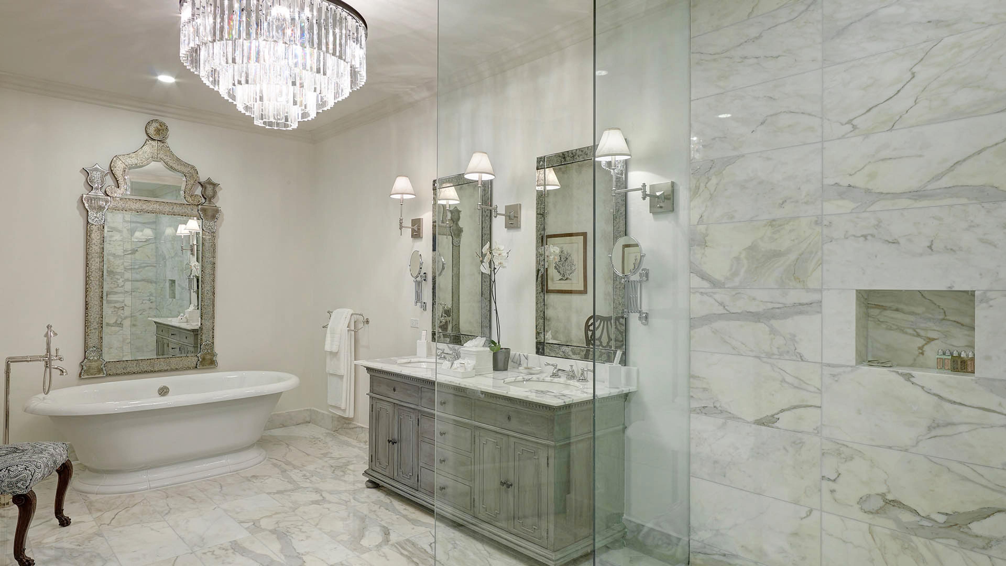 A bathroom with a freestanding tub, an ornate mirror, a crystal chandelier, a double vanity, and a glass-walled shower.