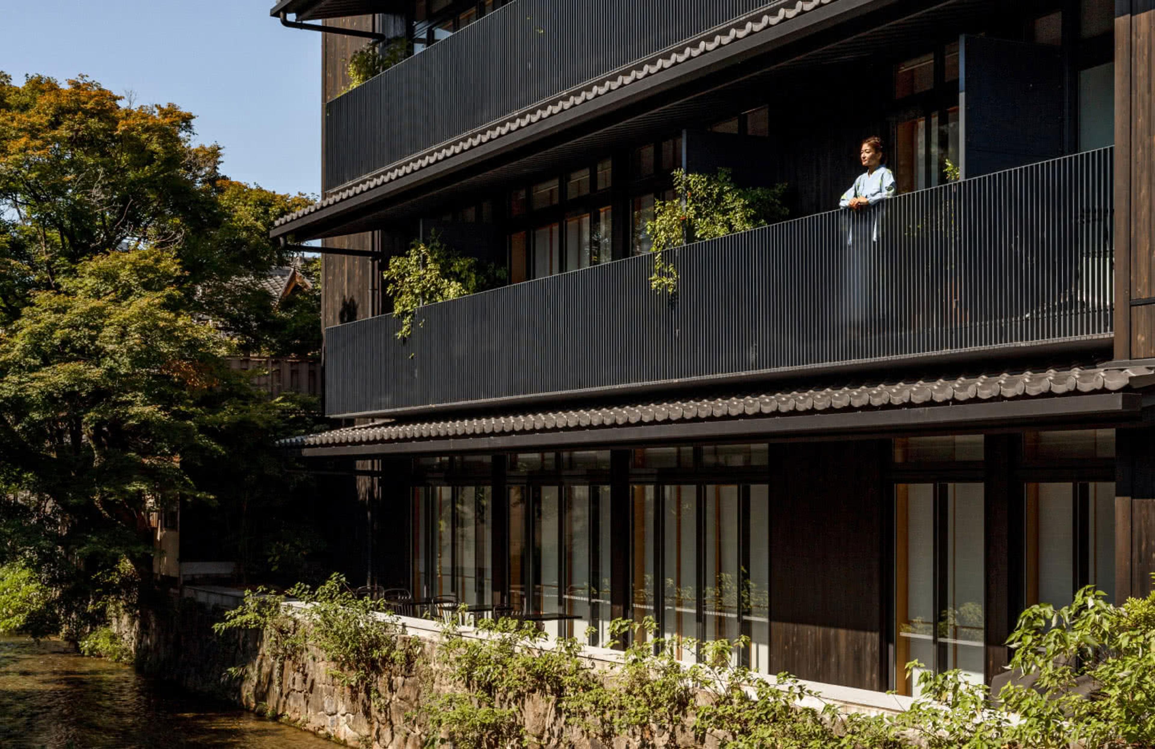 the hotel's dark timber upper floors and balconies seen across a tree-lined canal at dusk, with a robed figure standing at a distant balcony rail