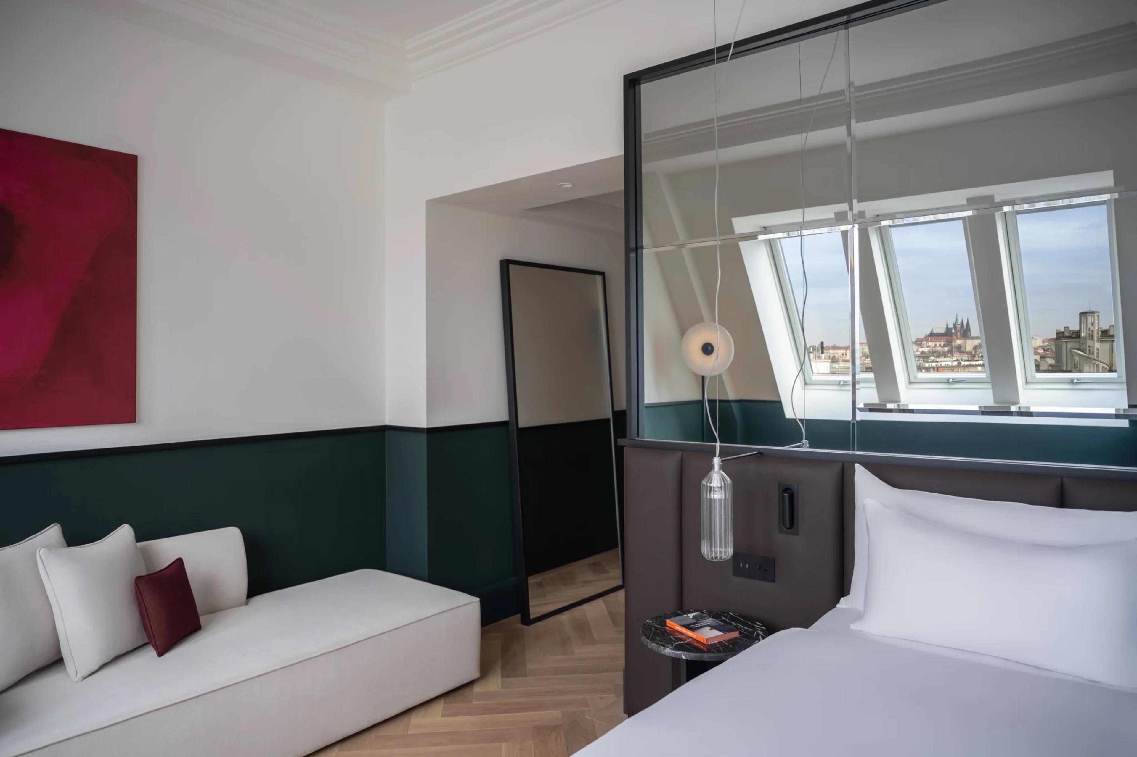 A bed beside a glass partition, with a window in the sloping roof showing the towers and rooftops of Prague Castle.