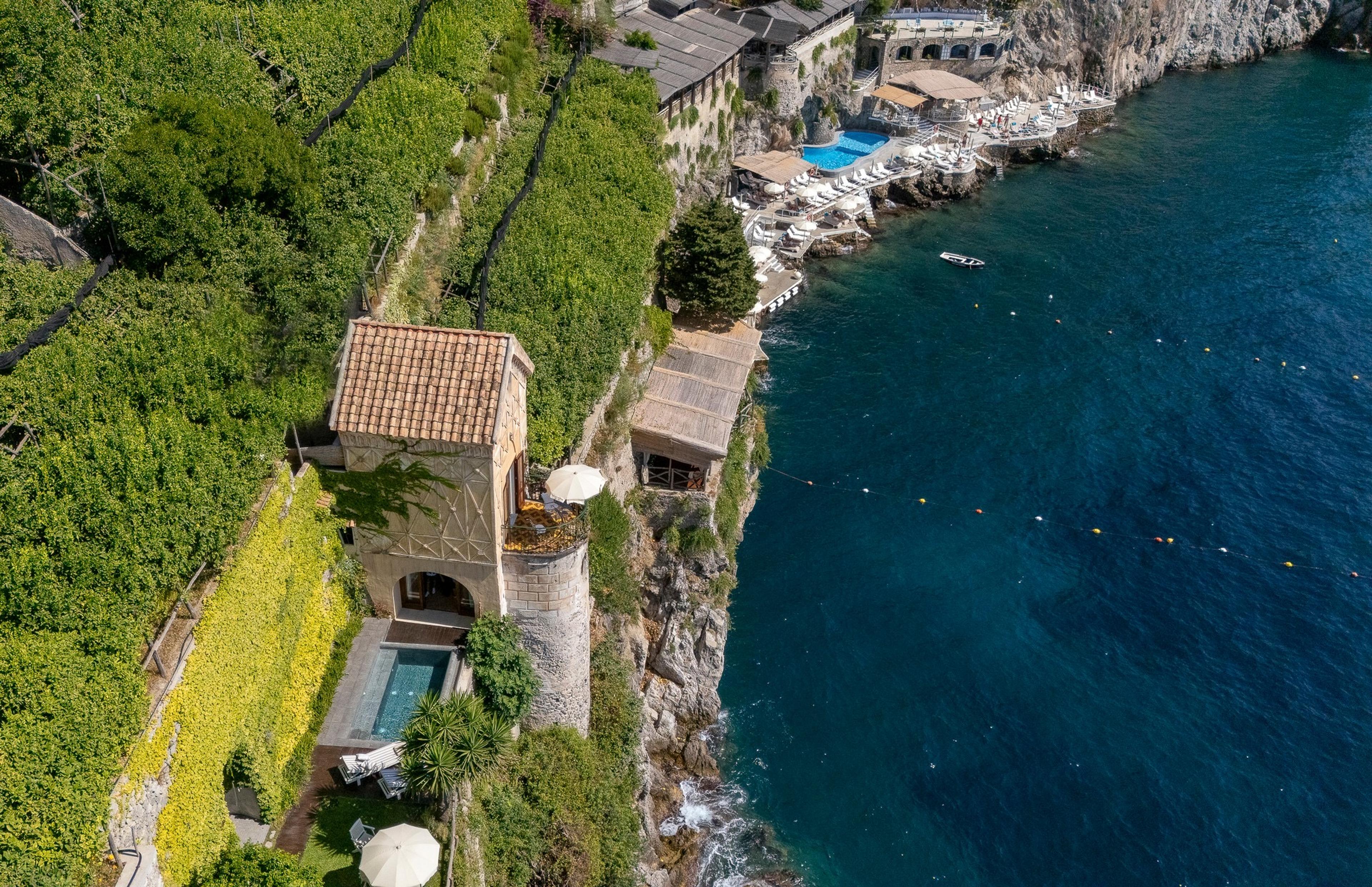 An aerial view of a hotel built into a cliffside, with two pools, sun loungers, a boat on the water, and a hillside town across the bay.