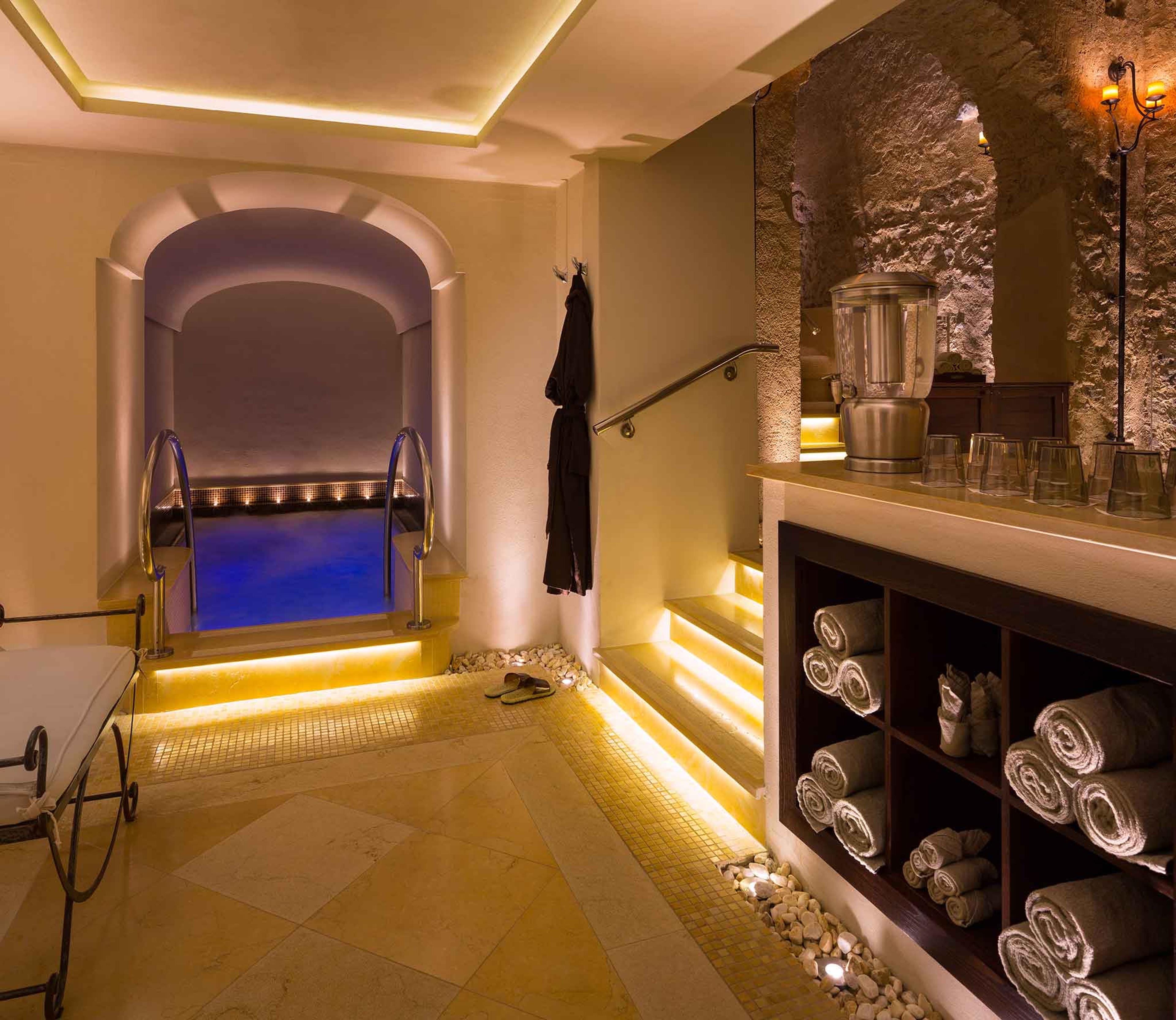 an indoor plunge pool lit in blue, reached by a few steps, next to a shelf of rolled towels and a robe hanging on the wall