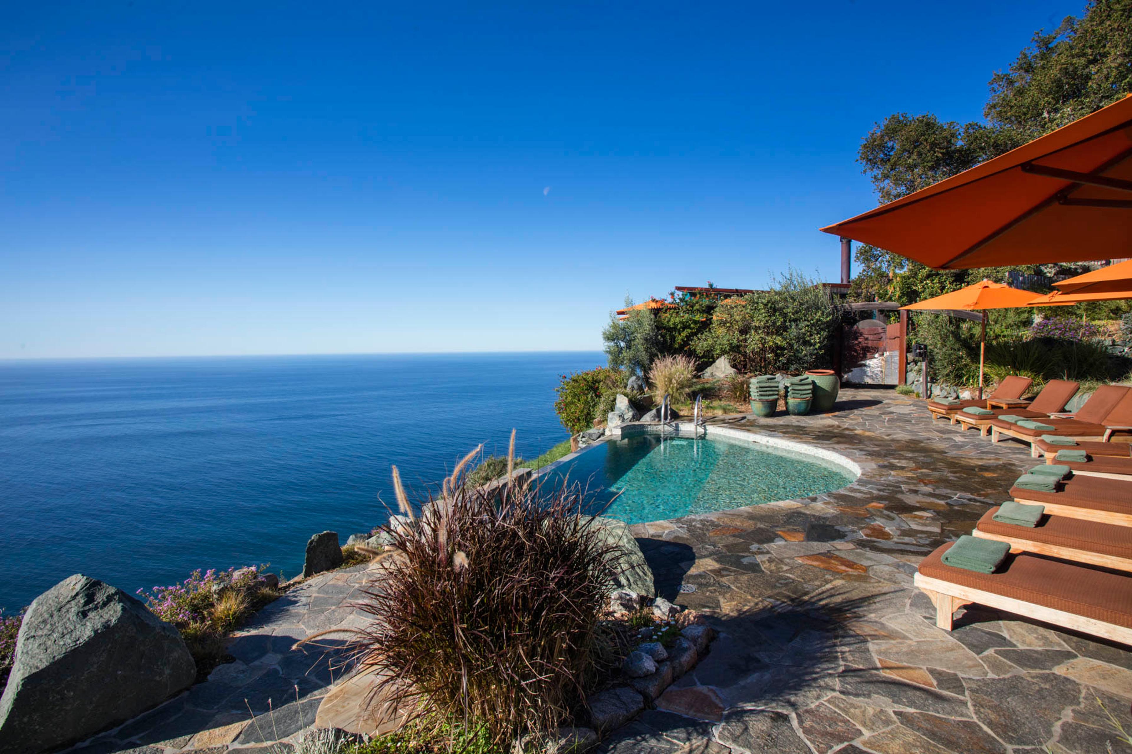 An infinity pool on a stone terrace overlooking the ocean, with loungers and orange umbrellas to one side and rocks and shrubs in the foreground.