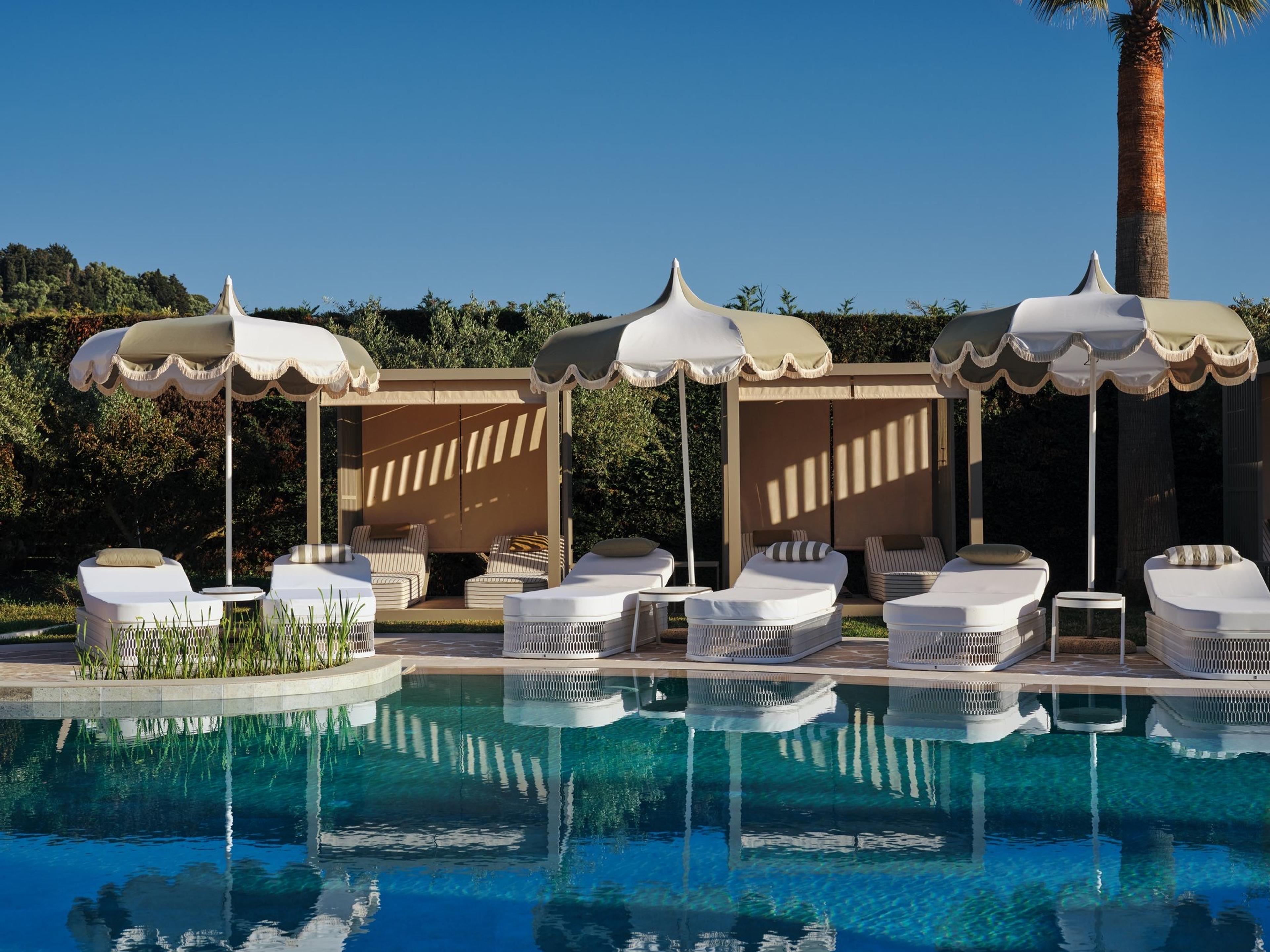 Three scallop-canopied daybeds beside a pool, with a palm tree and a clipped hedge behind them.