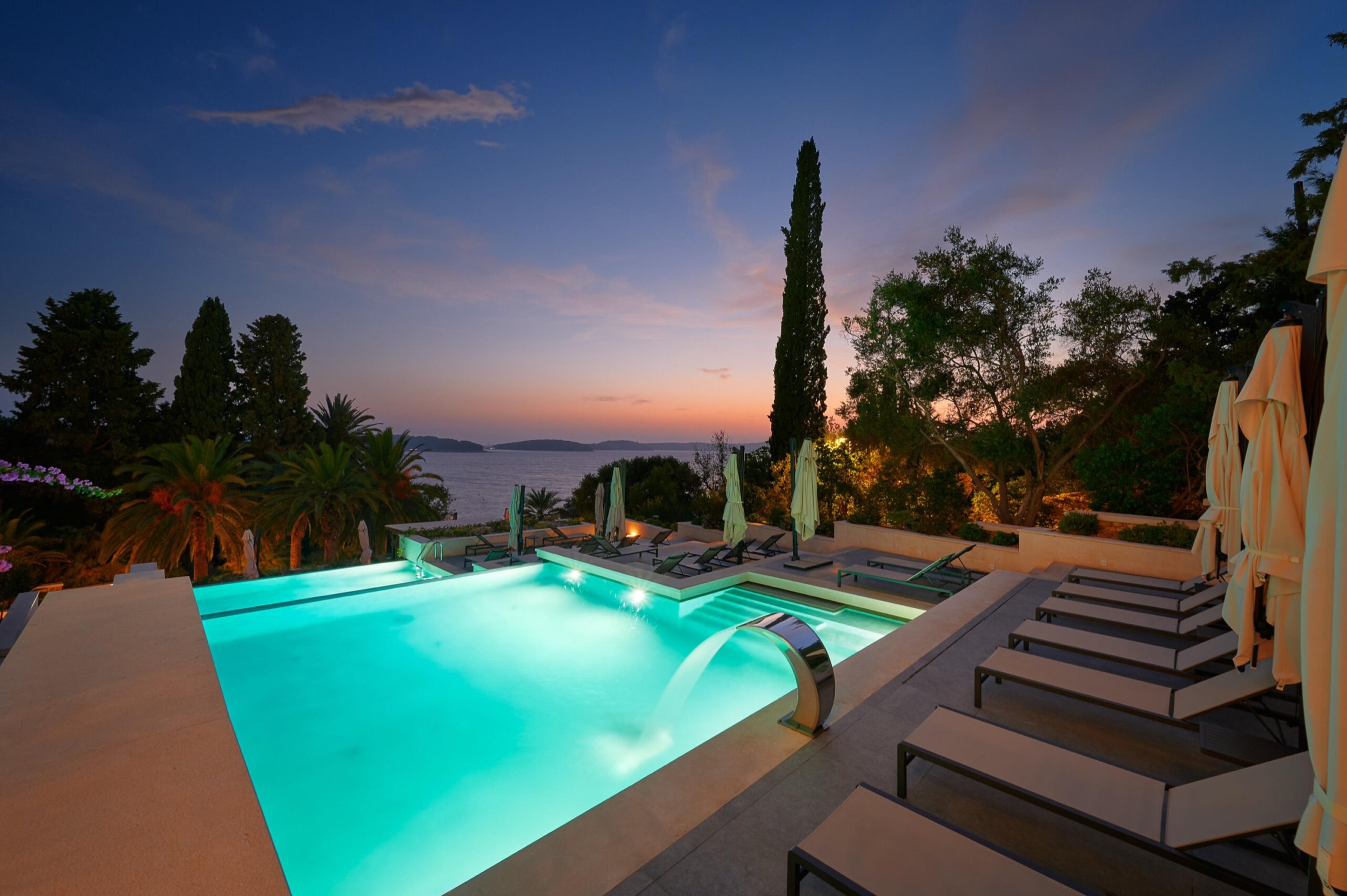 An outdoor pool lit turquoise at dusk, with sun loungers along one side and a view of the sea and pine-covered hills beyond.