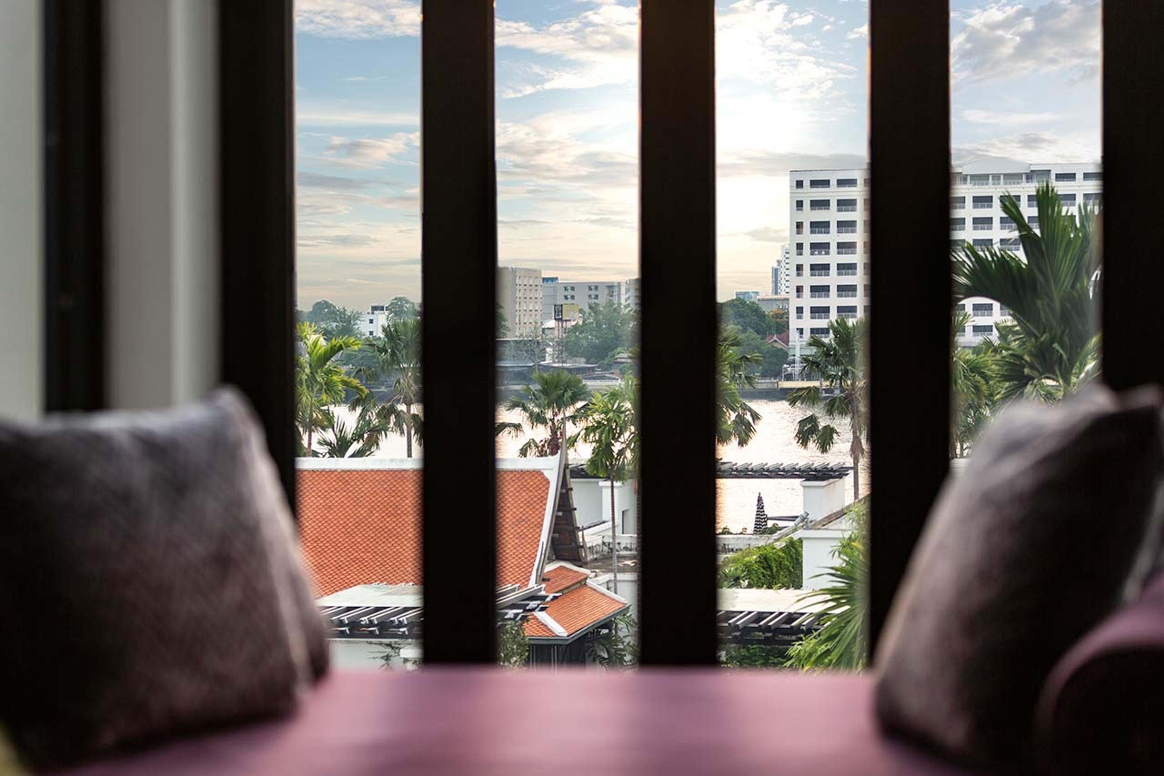 A view from a seating area past dark wooden shutters over orange-tiled temple roofs, palm trees and a river, with high-rise buildings on the far bank.