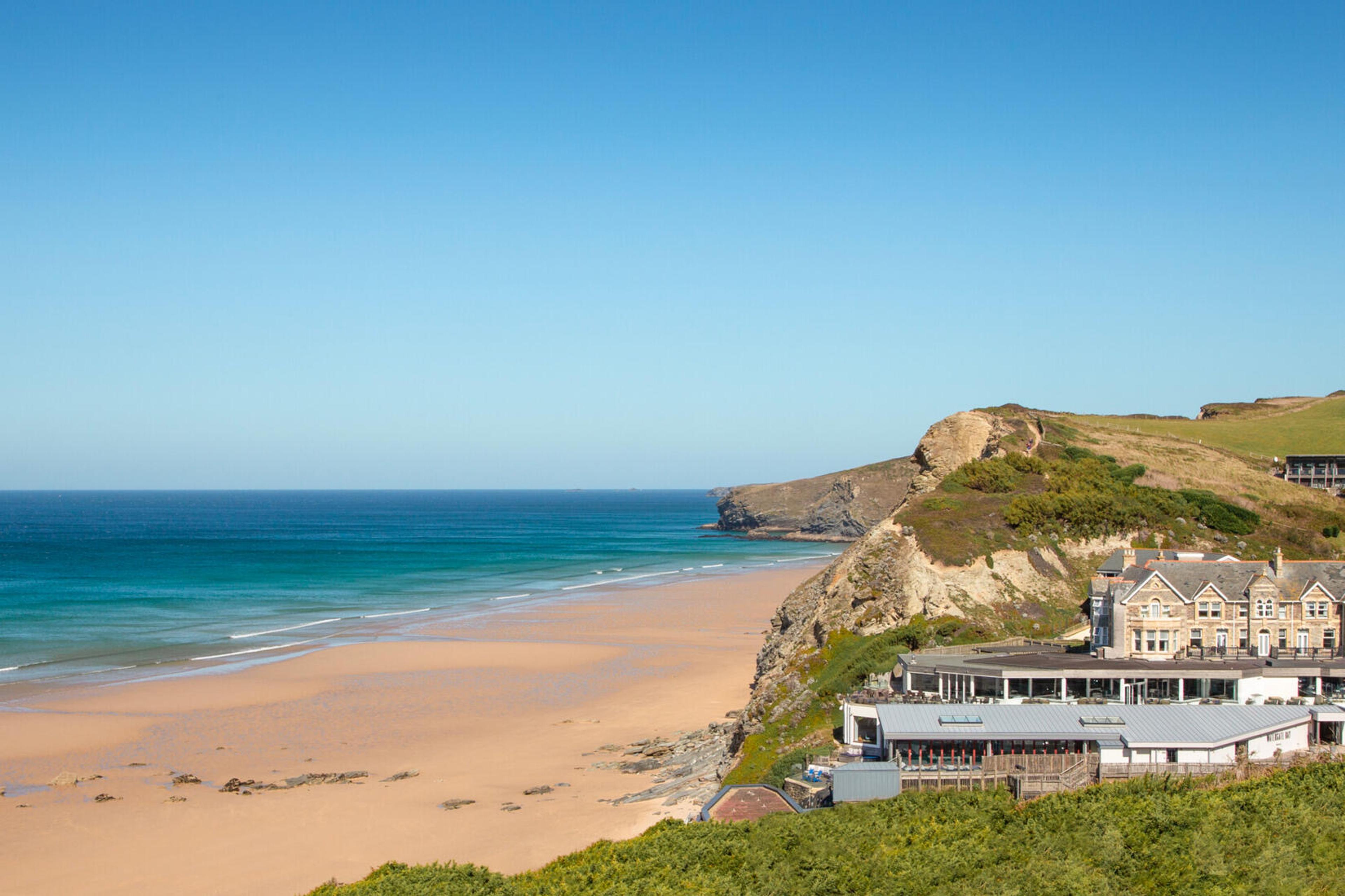 An aerial view of a stone hotel building on a headland above a sandy beach, with a terrace and outbuildings below it.