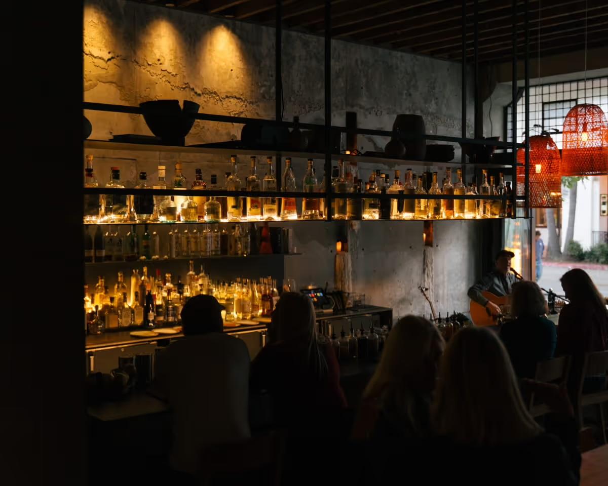 A dimly lit bar with backlit shelves of bottles against a rough concrete wall, red woven pendant lamps, and a row of seated guests with their backs to the camera facing a guitarist playing near the open doorway.