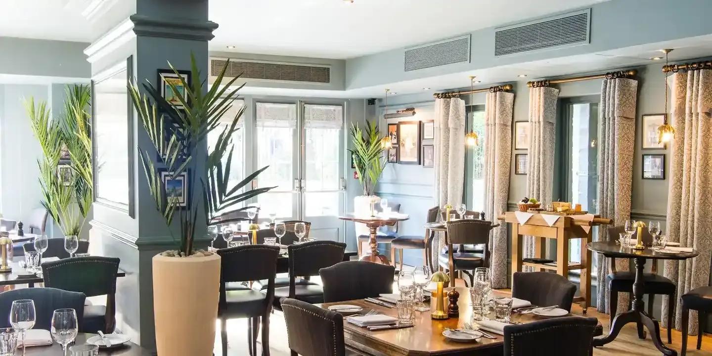 A restaurant dining room with pale blue walls, a tall palm in a pot beside a grey column, tables laid with white napkins, and tall windows with curtains at the back.