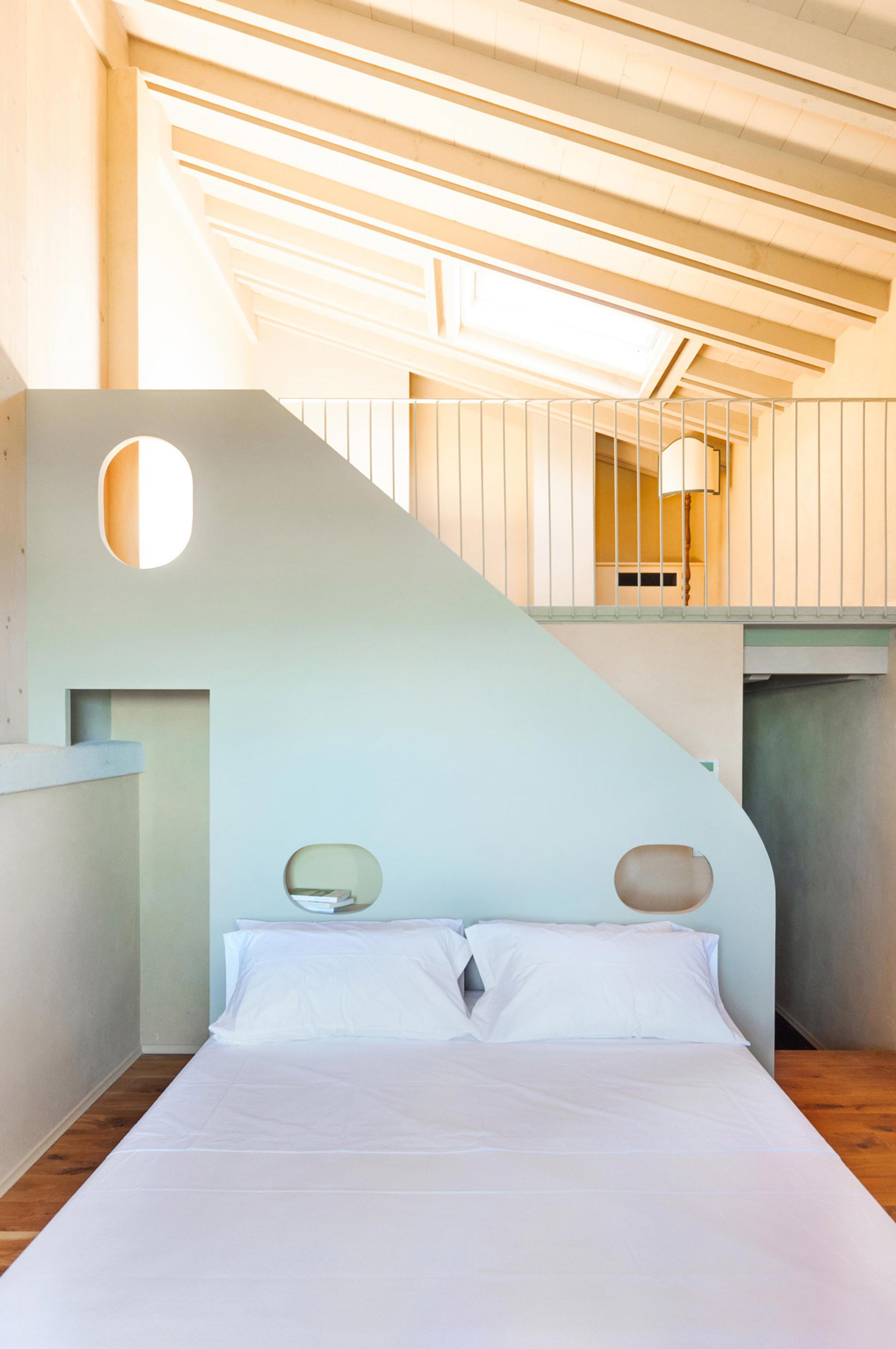 A bedroom with a curved pastel green wall holding oval cut-out shelves, a white-linened bed below and a mezzanine landing with a skylight above.