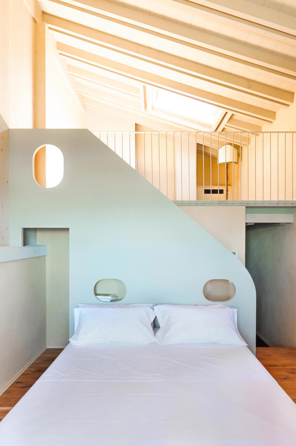 A bedroom with a curved pastel green wall holding oval cut-out shelves, a white-linened bed below and a mezzanine landing with a skylight above.