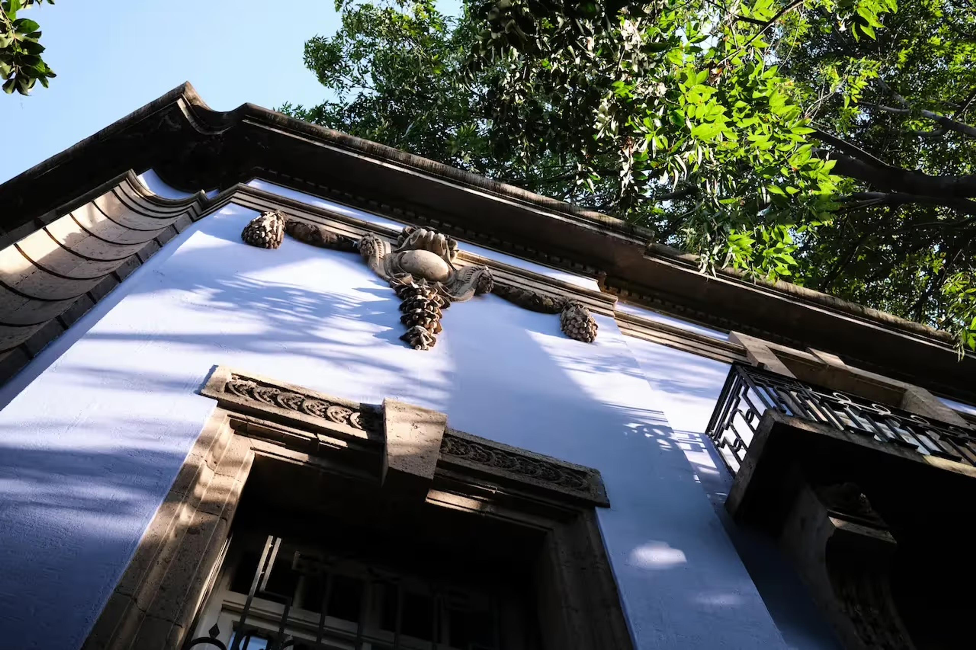 A carved stone pediment and roofline of a pale building rise under a canopy of trees, seen from below.