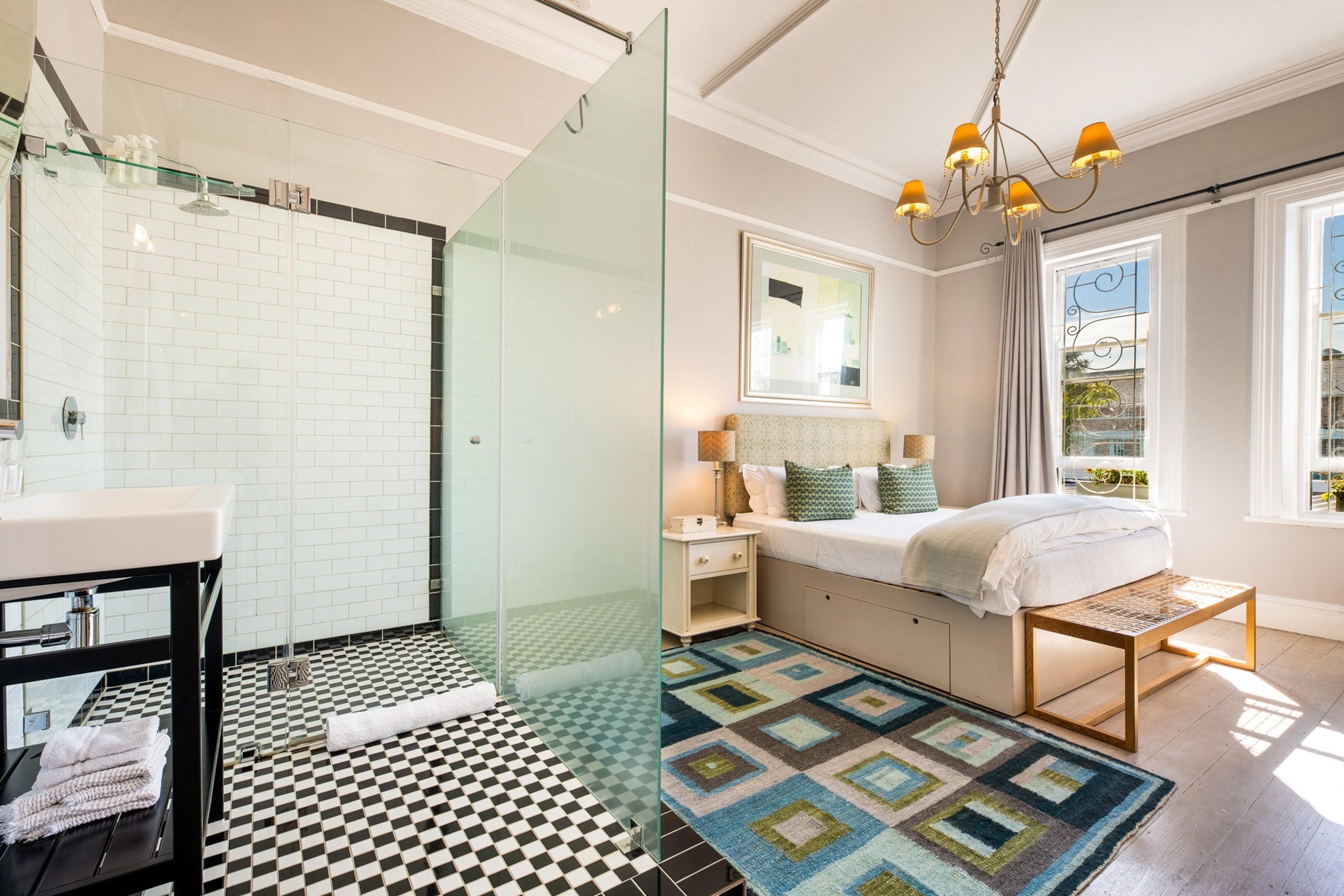 a bedroom with an open shower behind a glass partition, black and white checked floor tiles, and a patchwork rug beside the bed