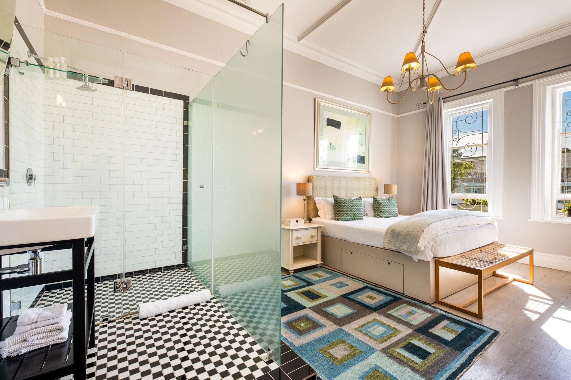 a bedroom with an open shower behind a glass partition, black and white checked floor tiles, and a patchwork rug beside the bed