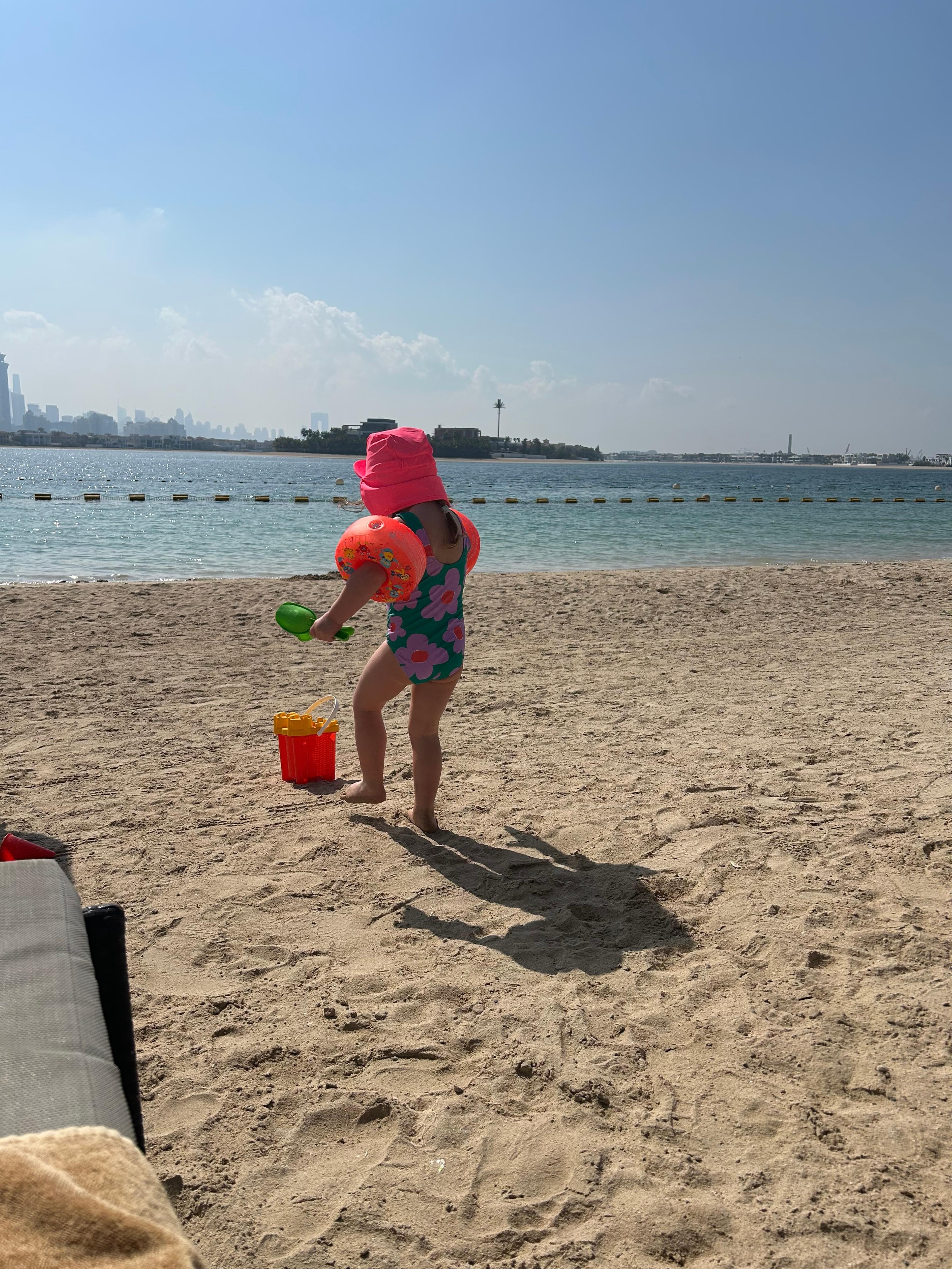 A child in a sunhat and armbands playing at the edge of the beach, the Dubai Marina skyline across the water