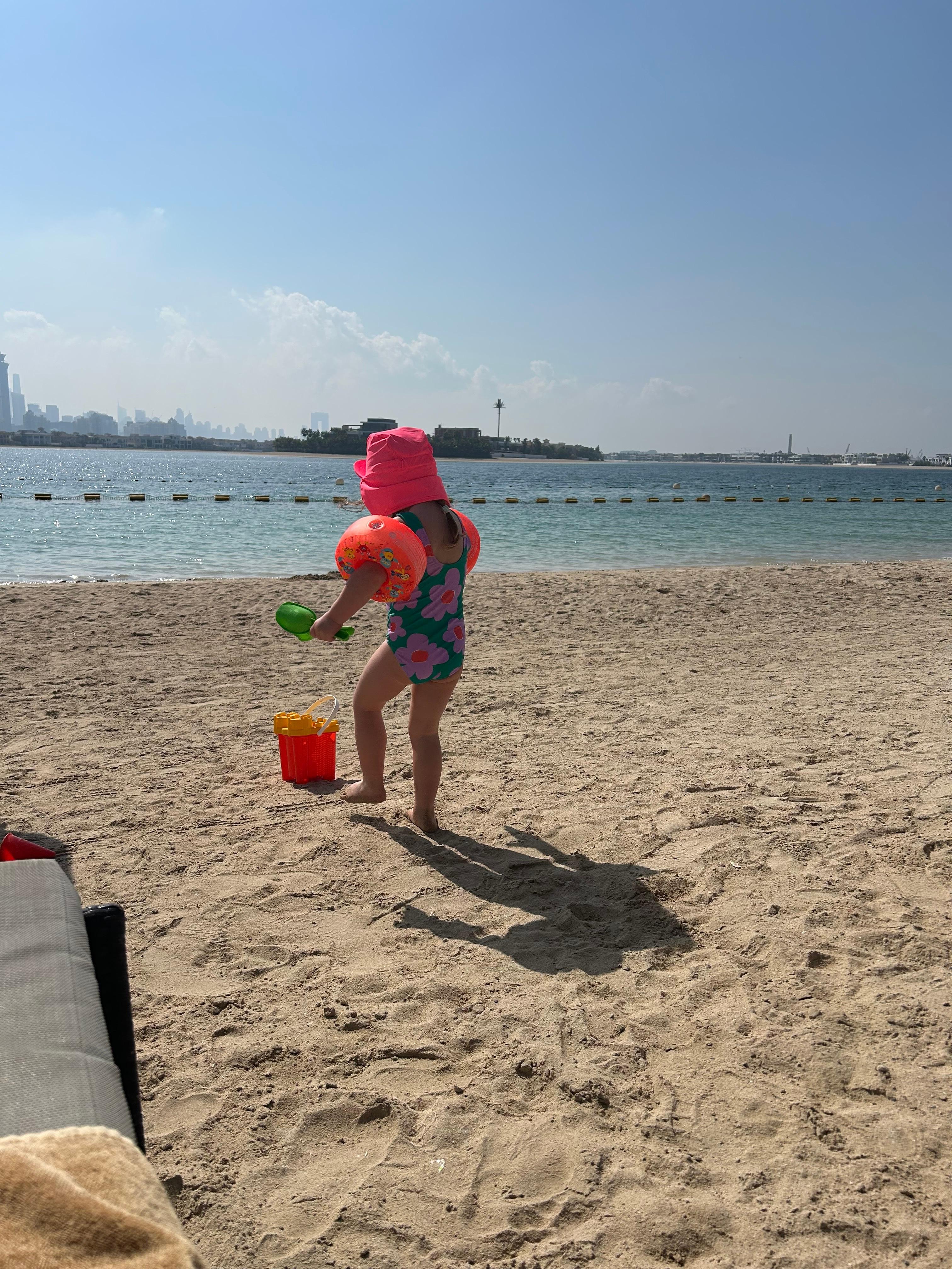 A child in a sunhat and armbands playing at the edge of the beach, the Dubai Marina skyline across the water