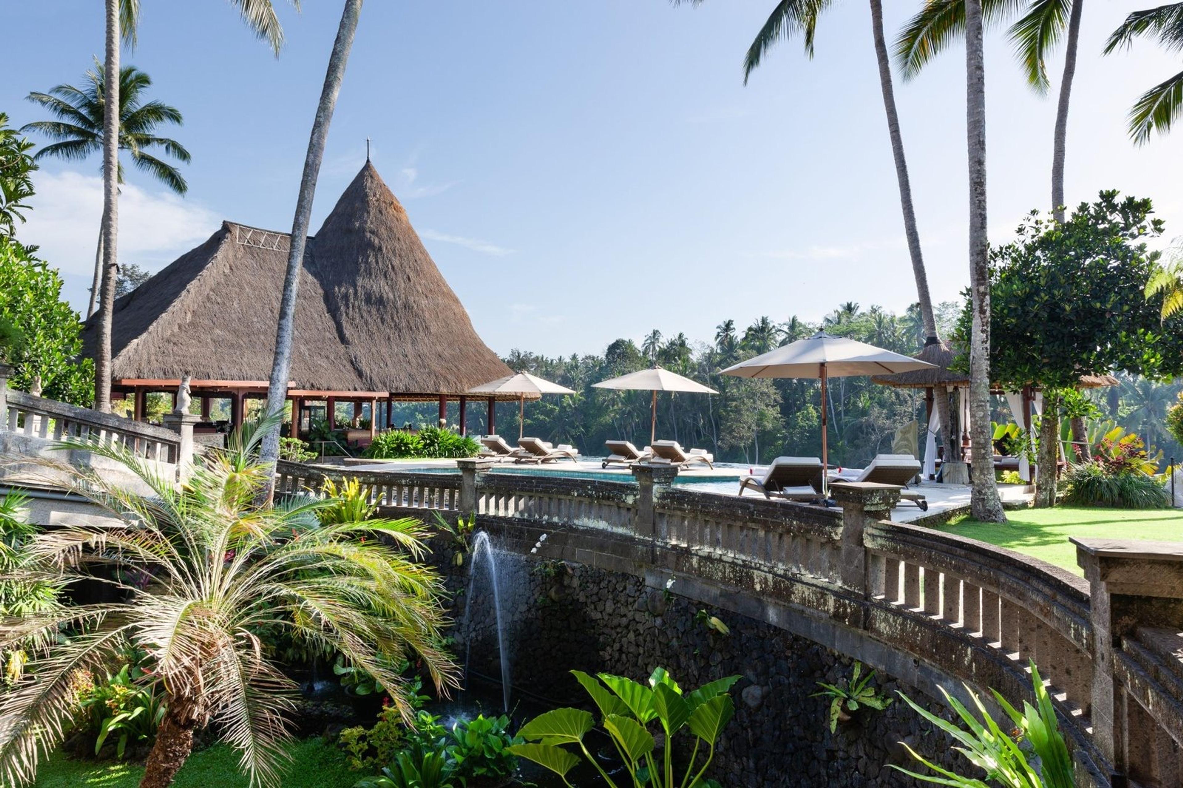 A stone bridge with a fountain crosses a pond toward a steep thatched pavilion, with a pool and sunloungers visible beyond it among palm trees.