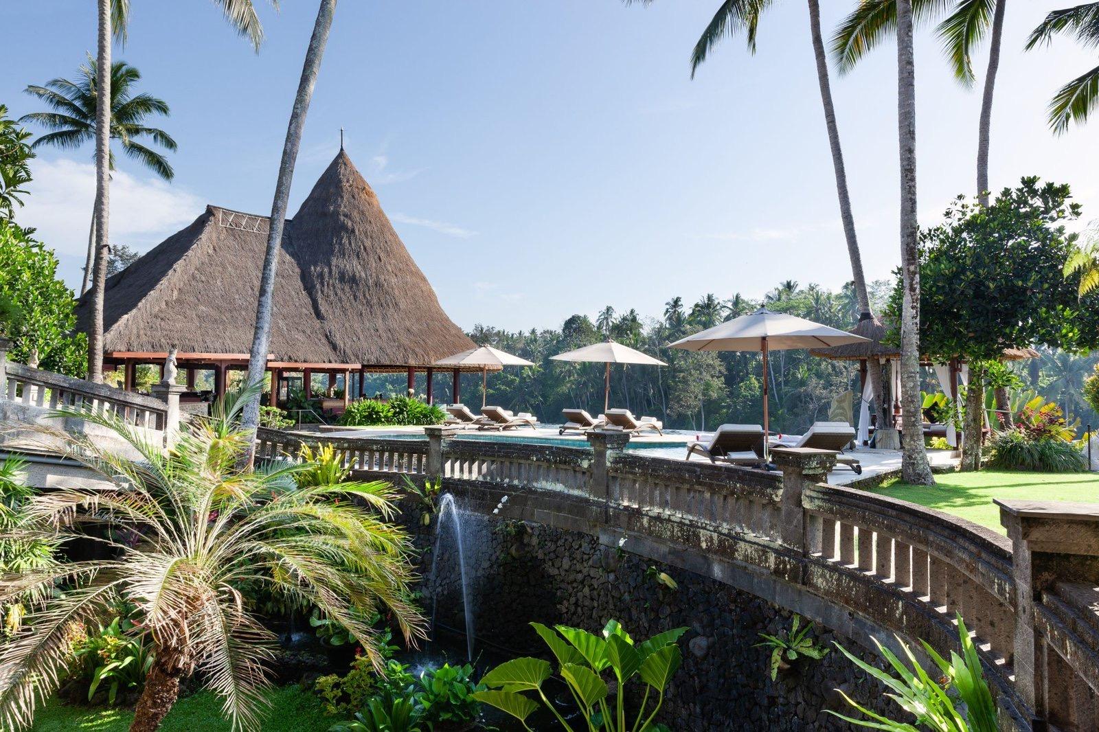 A stone bridge with a fountain crosses a pond toward a steep thatched pavilion, with a pool and sunloungers visible beyond it among palm trees.