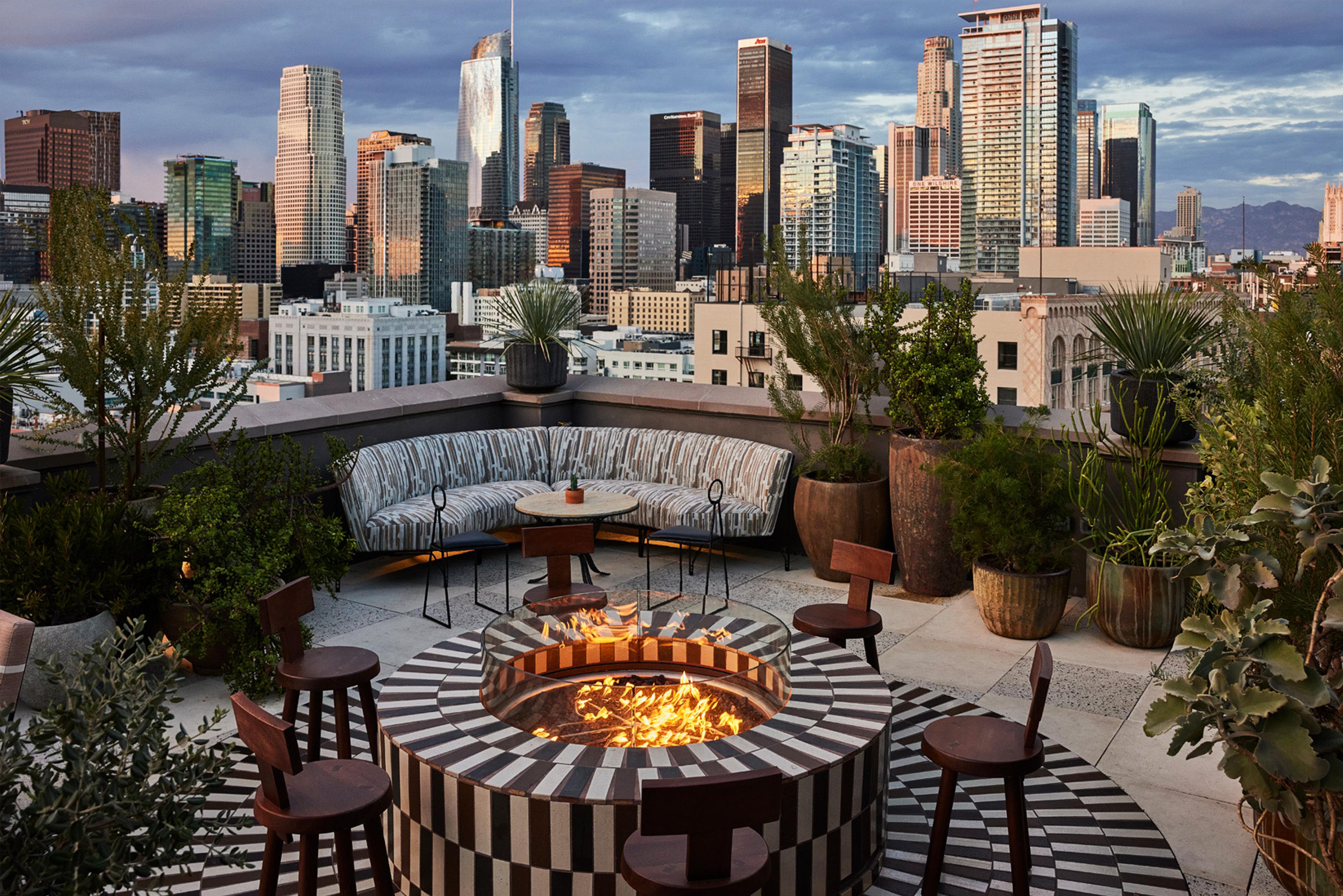 A rooftop lounge at dusk with couches around a round tiled fire pit, bar stools, potted plants, and the downtown Los Angeles skyline lit up behind.