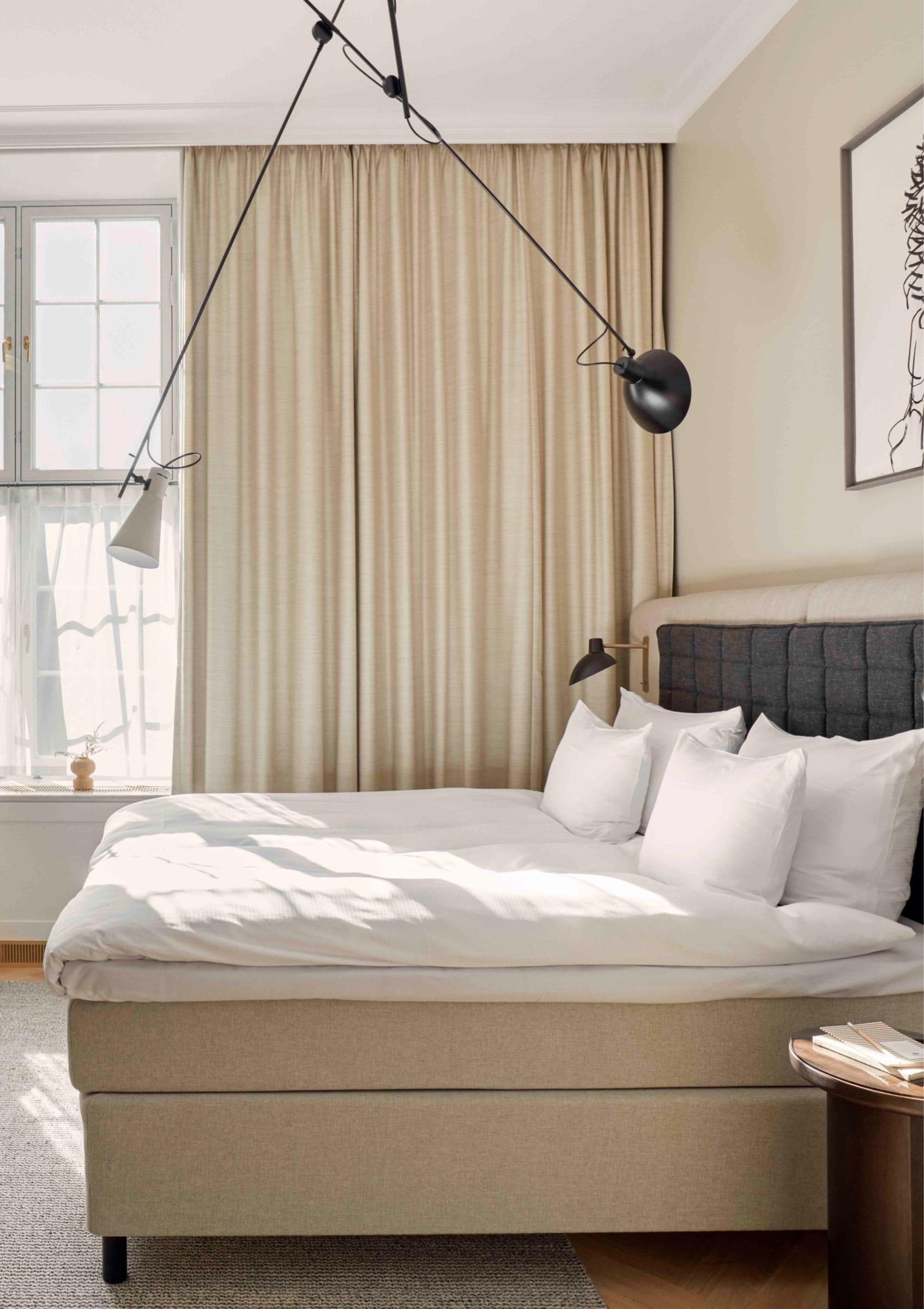 A bedroom with a bed in white linen against a dark tufted headboard, two black pendant lamps hanging on crossed cords from the ceiling, and a curtained window to one side.