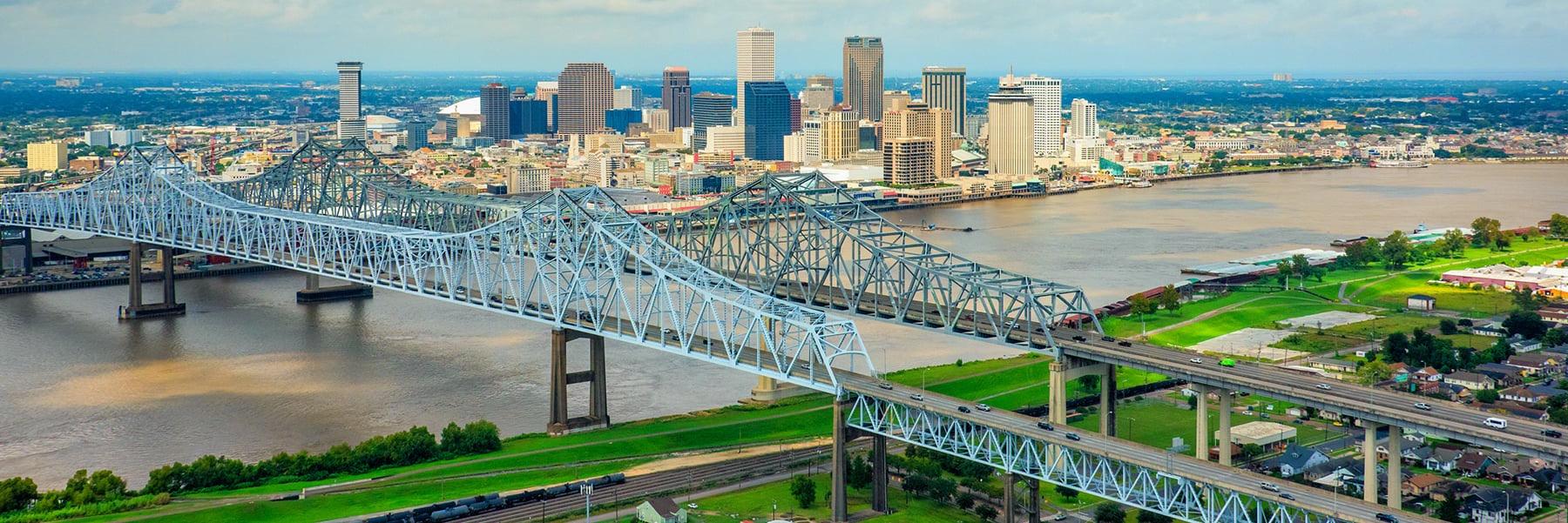 An aerial view of New Orleans showing a bridge crossing the Mississippi River with the downtown skyline beyond.