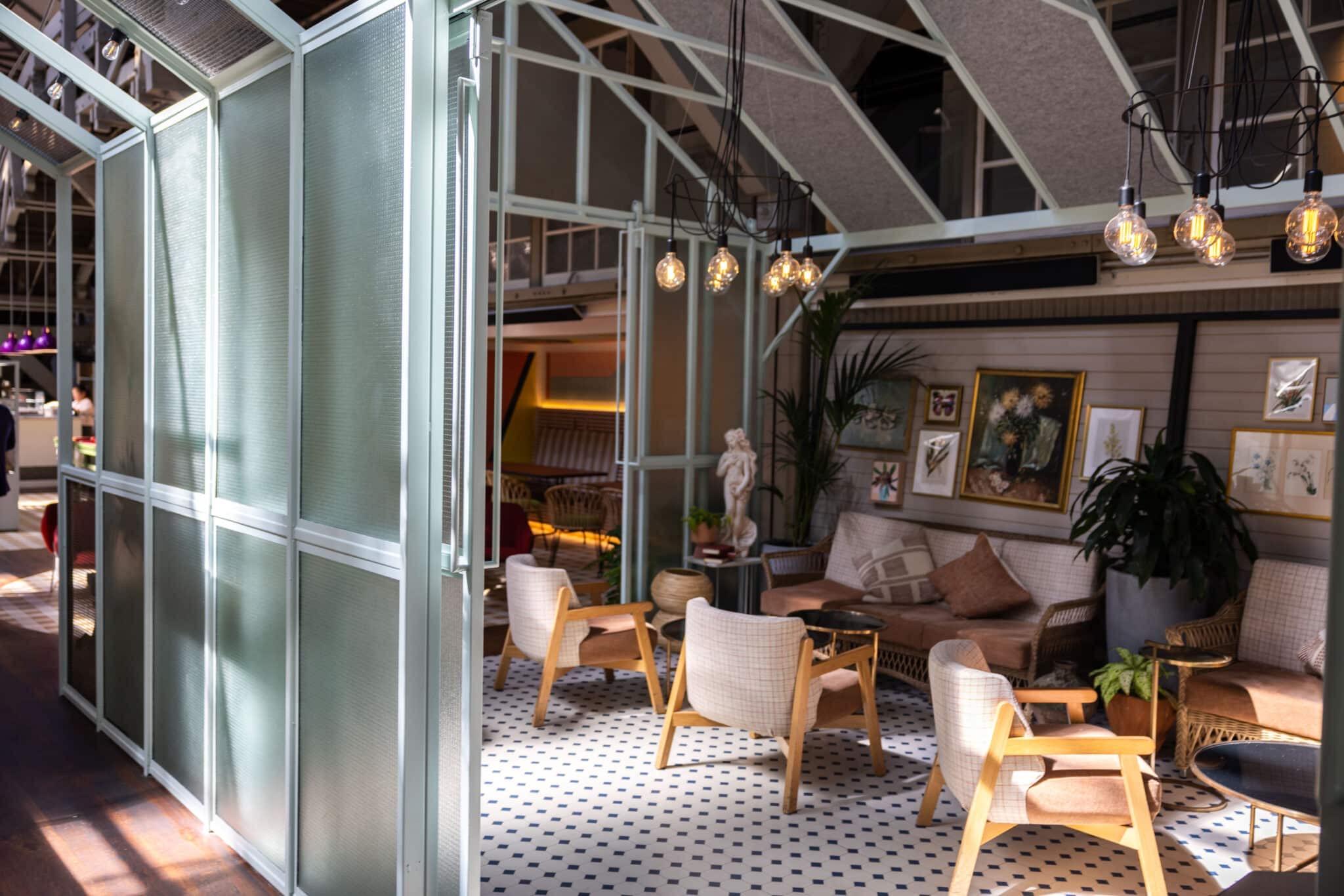 A lounge corner with checked armchairs, a glass partition wall, hanging bulb lights and framed botanical prints, with a person blurred in the background.
