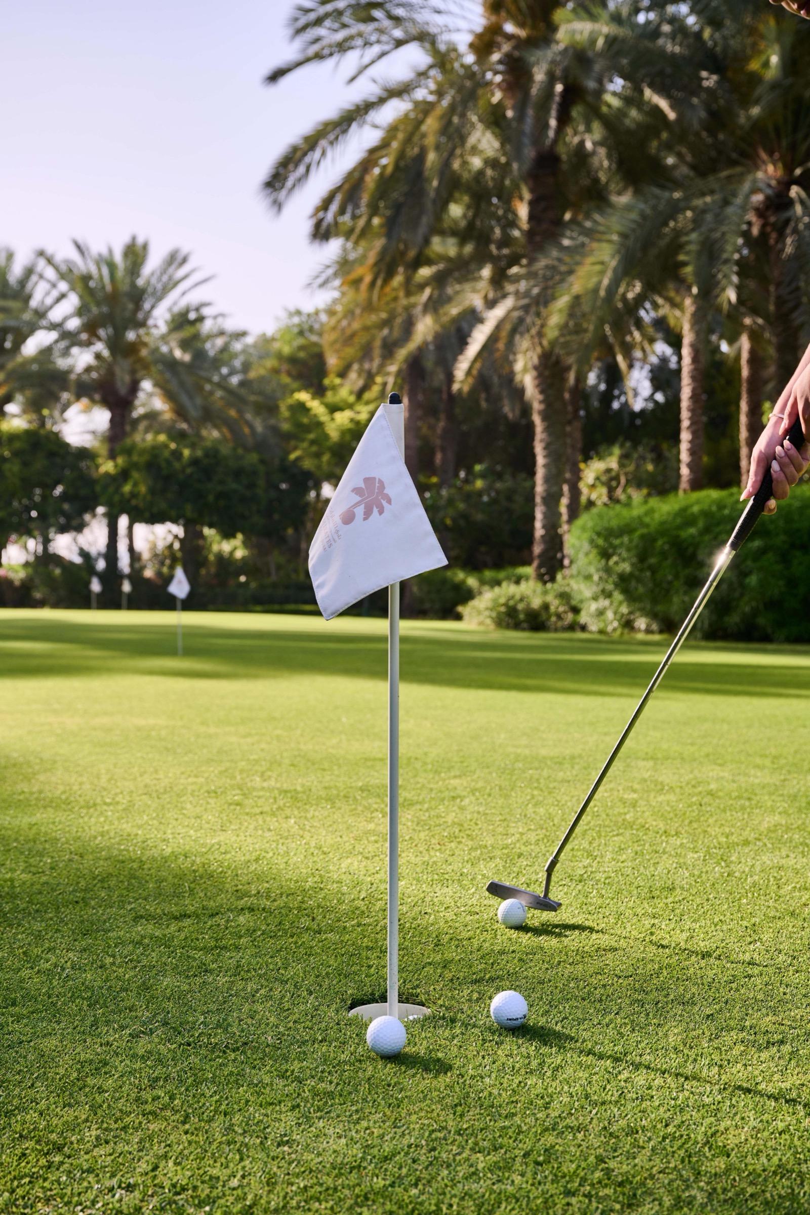 A putter and three golf balls sit on a green beside a flagstick printed with a small palm tree, palm trees behind.