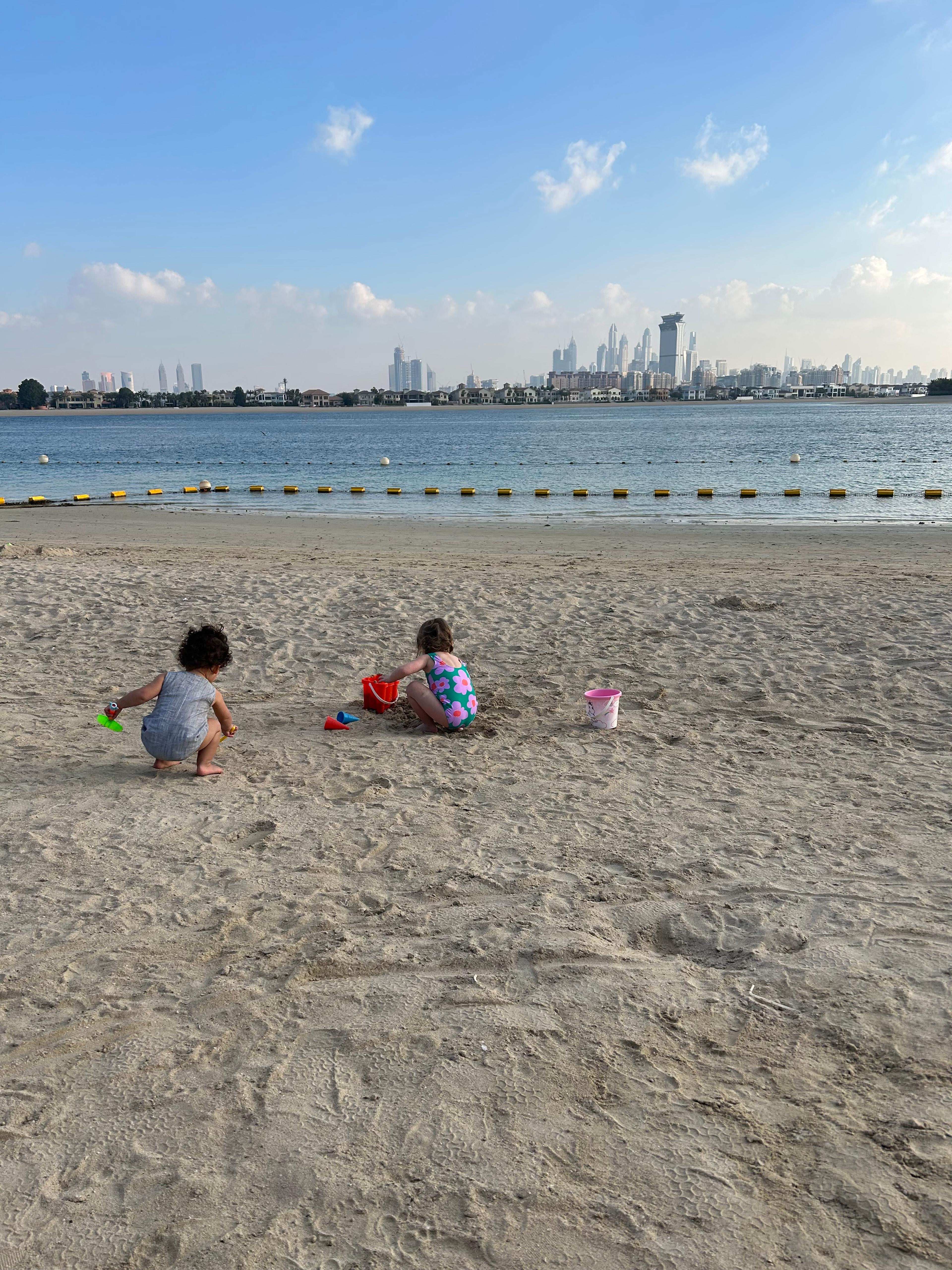 Two children playing in the sand with buckets, the sea and Dubai skyline behind them