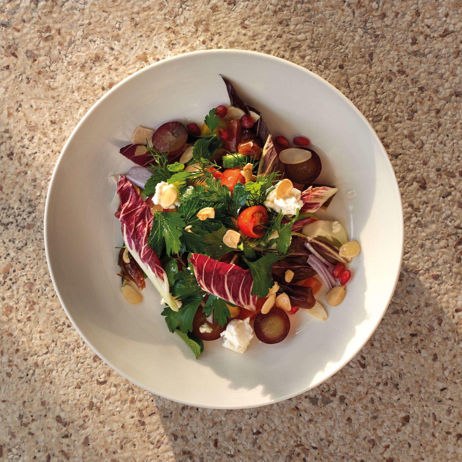 A white bowl holding a salad of radicchio, herbs, tomatoes, grapes and goat's cheese, on a speckled stone counter.