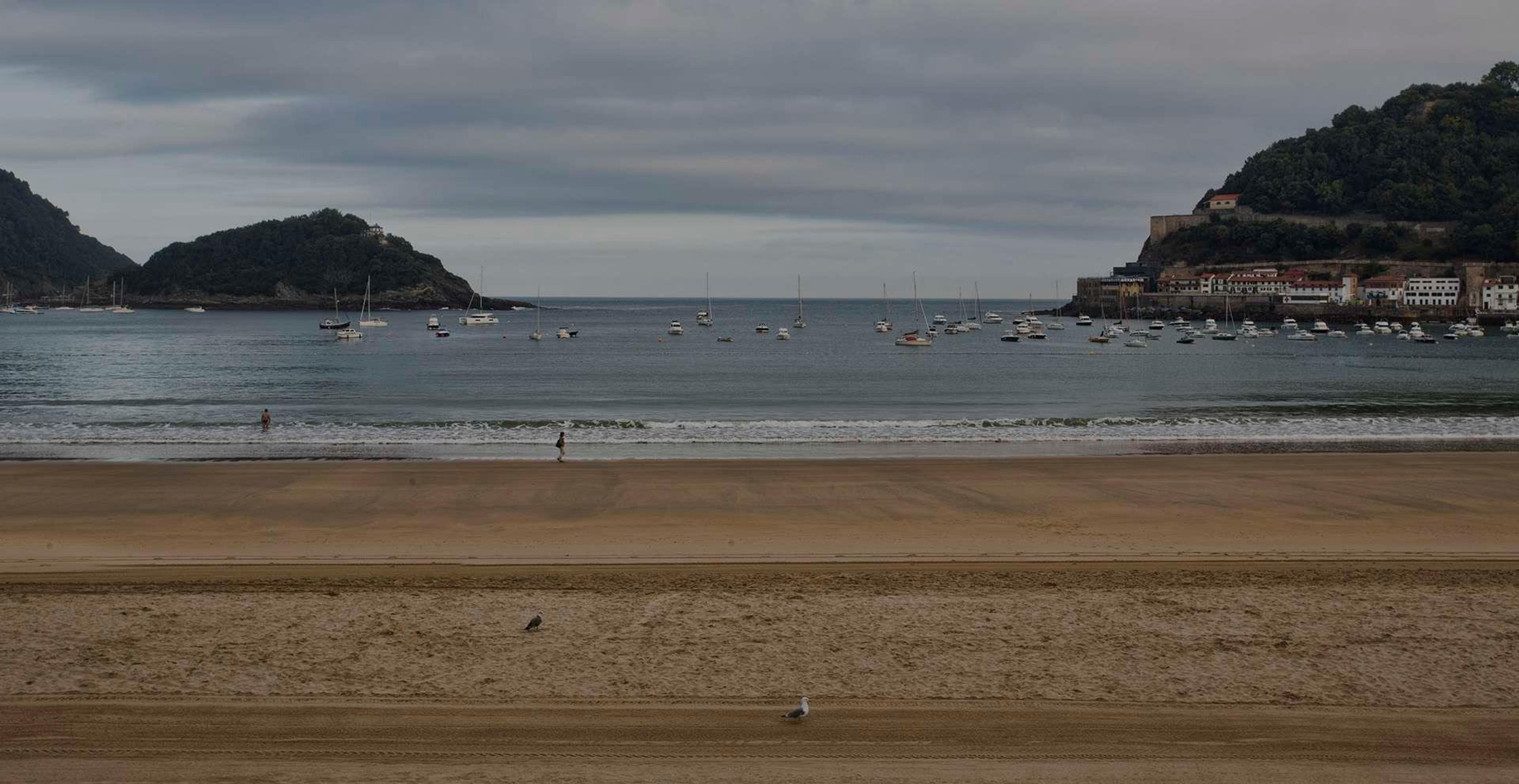 A wide, overcast view of La Concha beach with sailboats anchored in the bay and a wooded headland on either side.