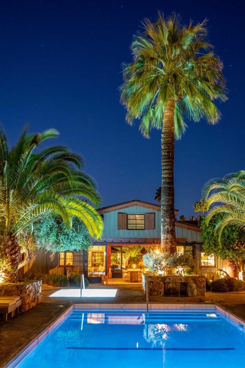 A two-storey wooden house with lit windows and a tall palm tree beside it, seen across a lit swimming pool at night.