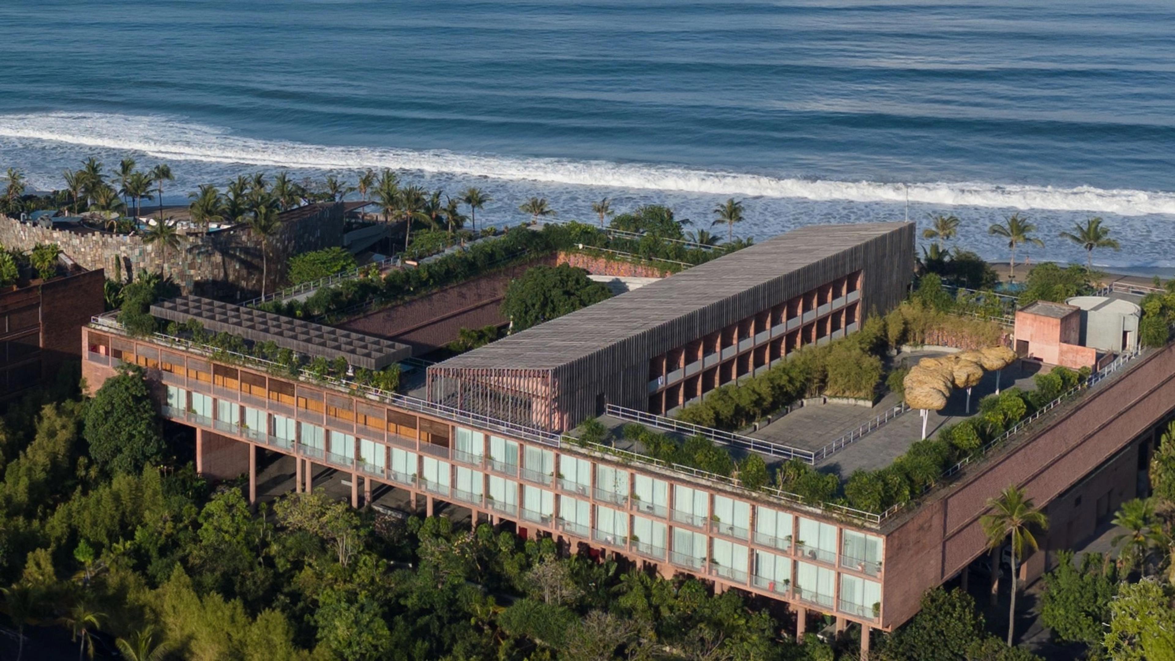 An aerial view of the hotel's long building along the shoreline, with guest corridors, a rooftop deck and solar panels between the beach and dense palm trees.