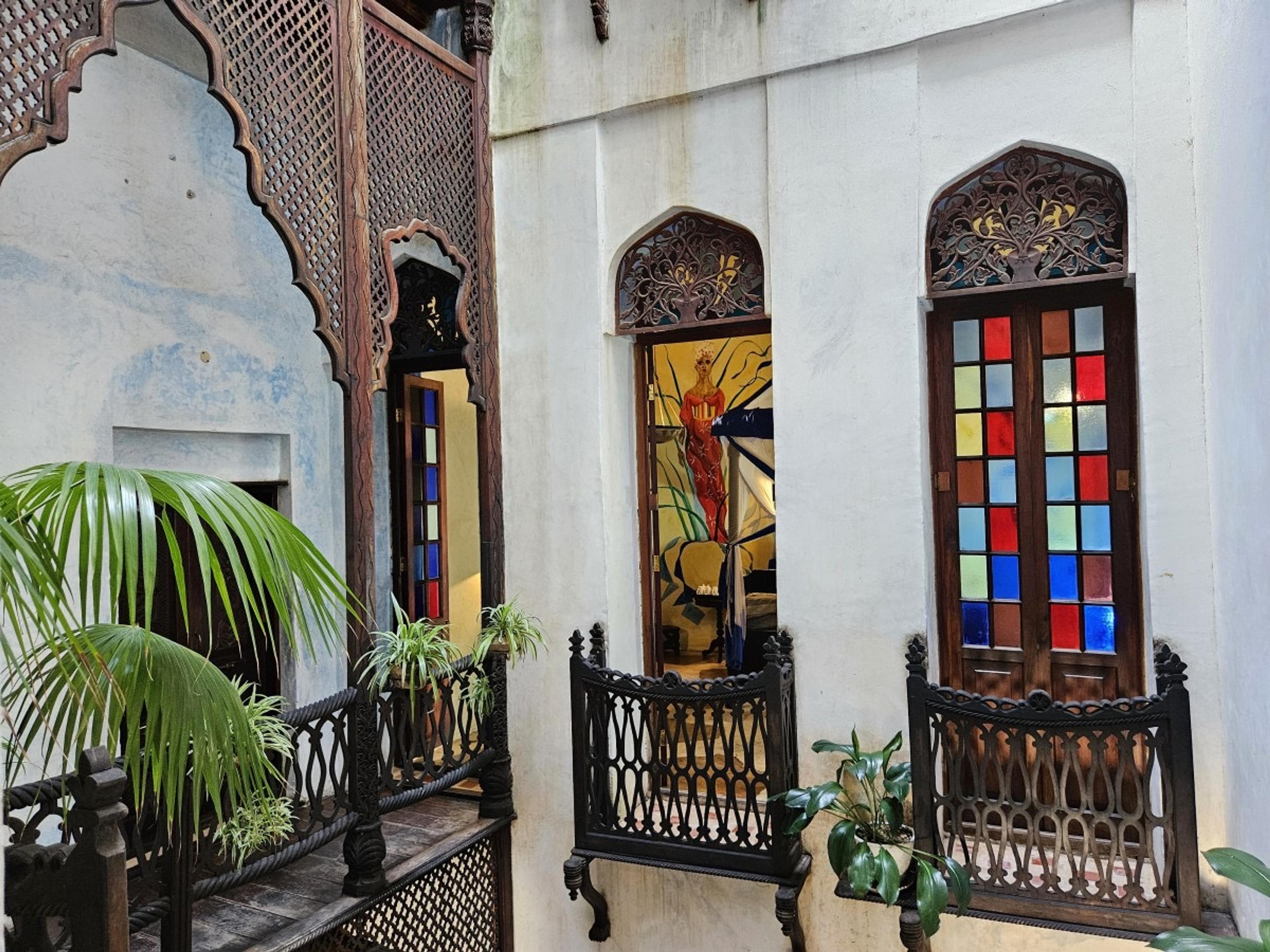 a wrought-iron balcony with potted palms on a white building facade, a window open onto a room painted with a mural of a robed figure