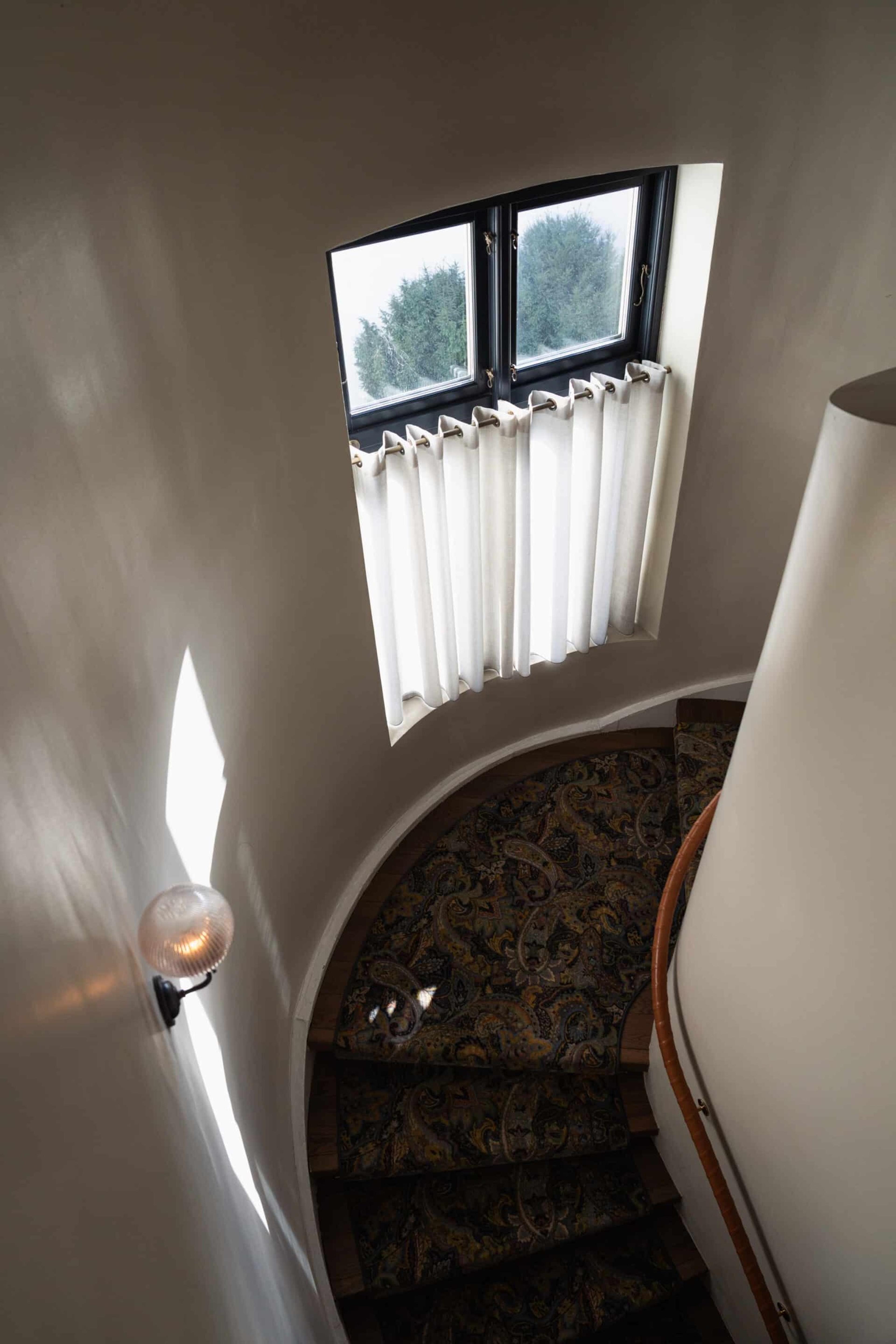 A curved staircase seen from above, lit by a small arched window with white curtains and a wall lamp, the stairs carpeted in a dark paisley runner.