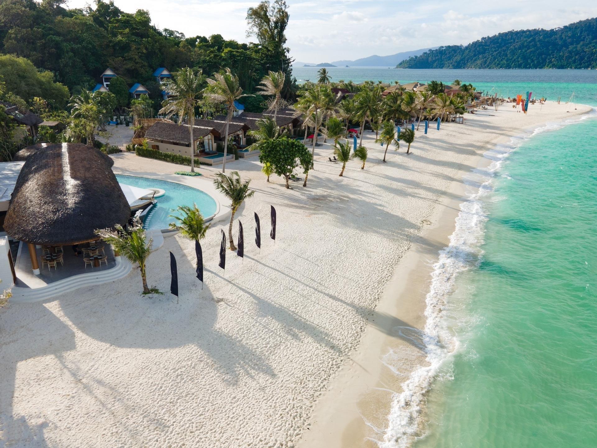 an aerial view of a thatched beach restaurant and pool beside a line of palm trees on a white sand beach, with forested hills and the sea beyond