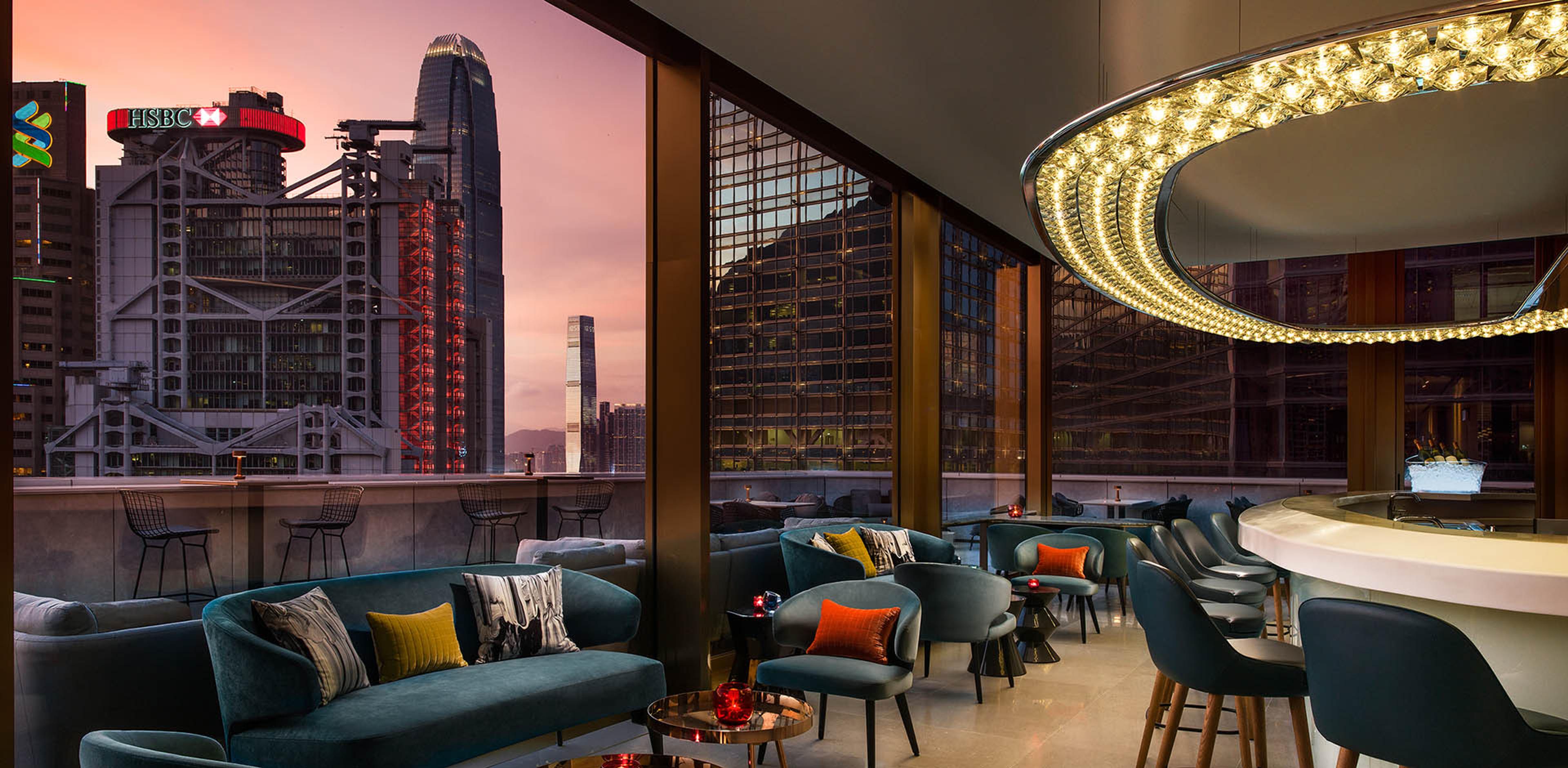 a rooftop bar at dusk with low sofas and barstools looking out over a lit skyline including the HSBC building