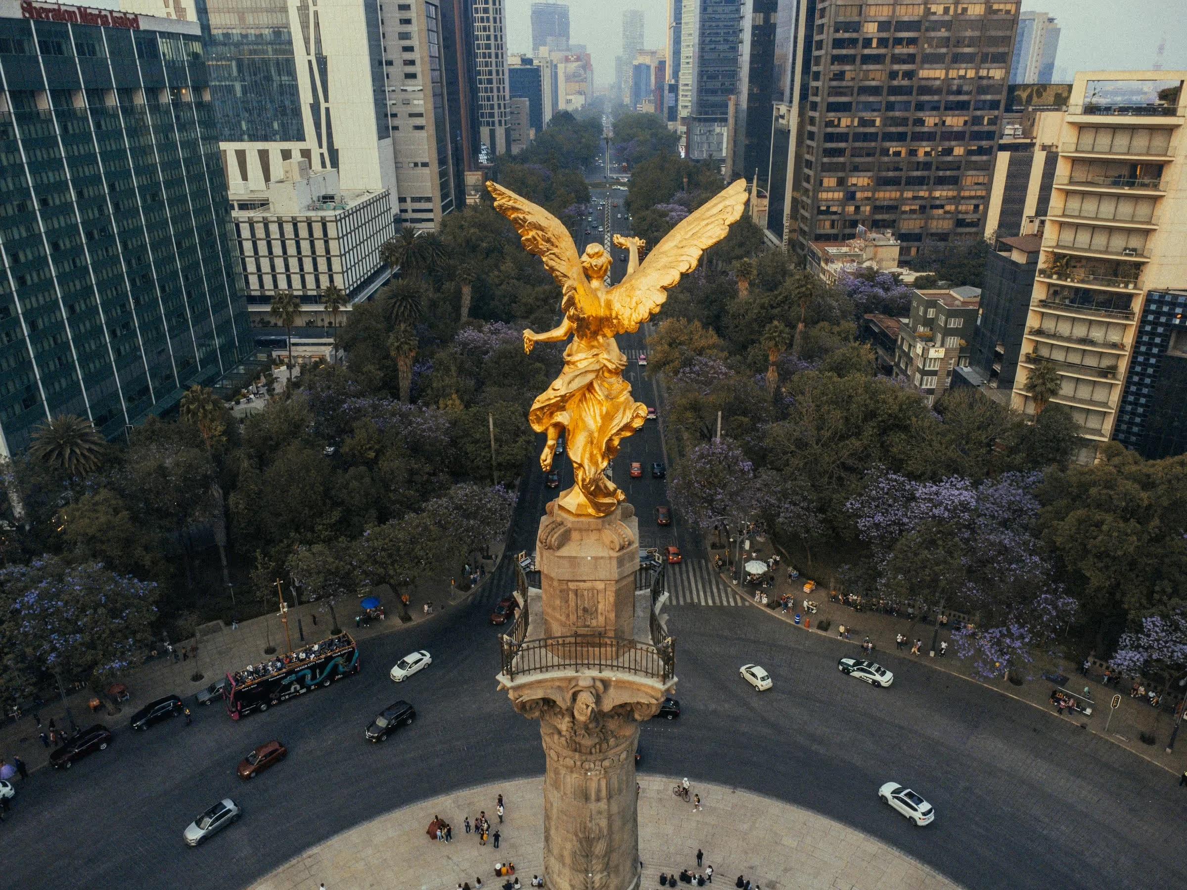 An aerial view of a golden winged angel statue atop its column on a Mexico City avenue, traffic circling below among jacaranda trees.