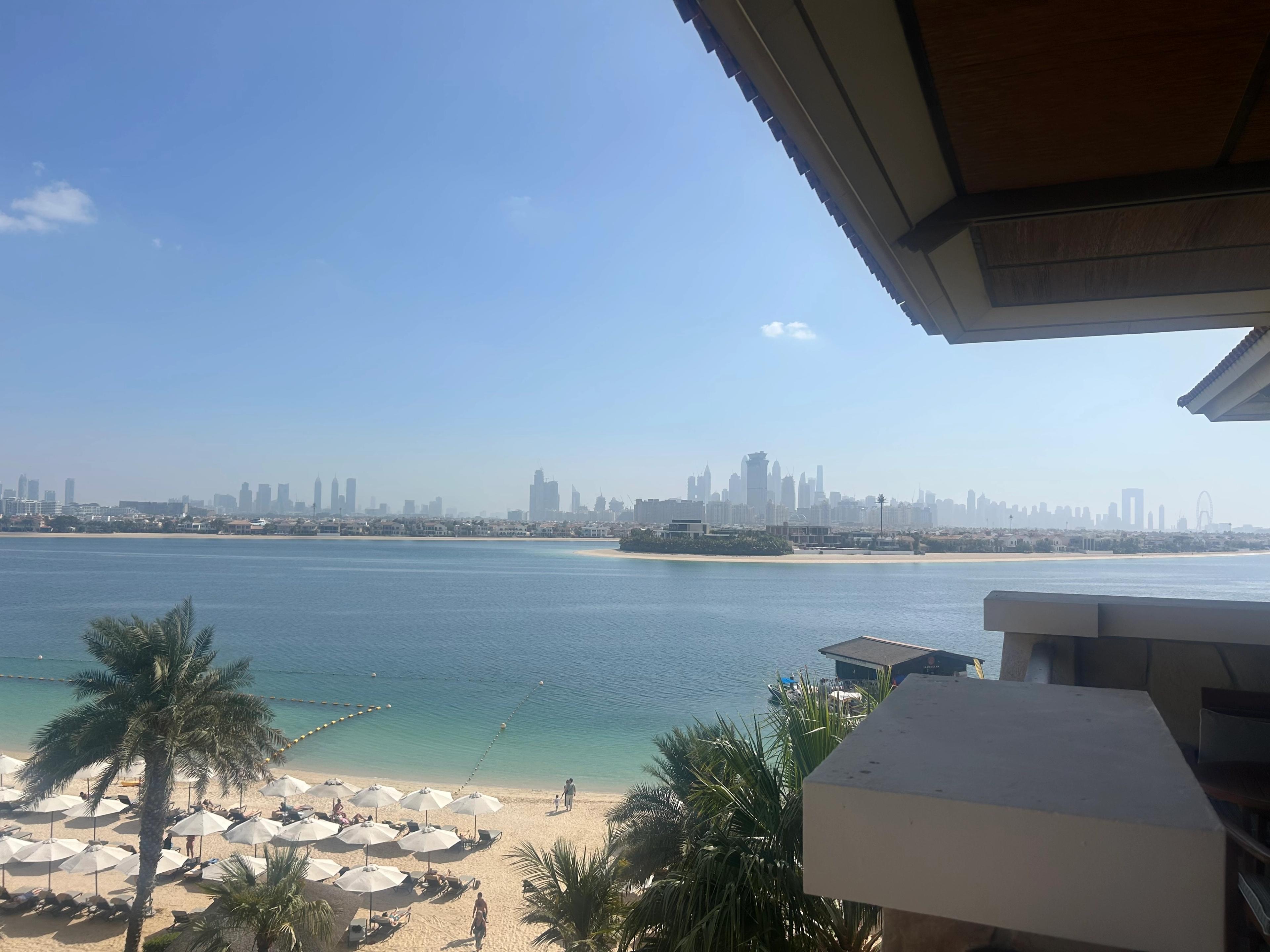 View from a hotel balcony under a red-tiled eave, over a beach of white umbrellas to the lagoon and Dubai Marina skyline
