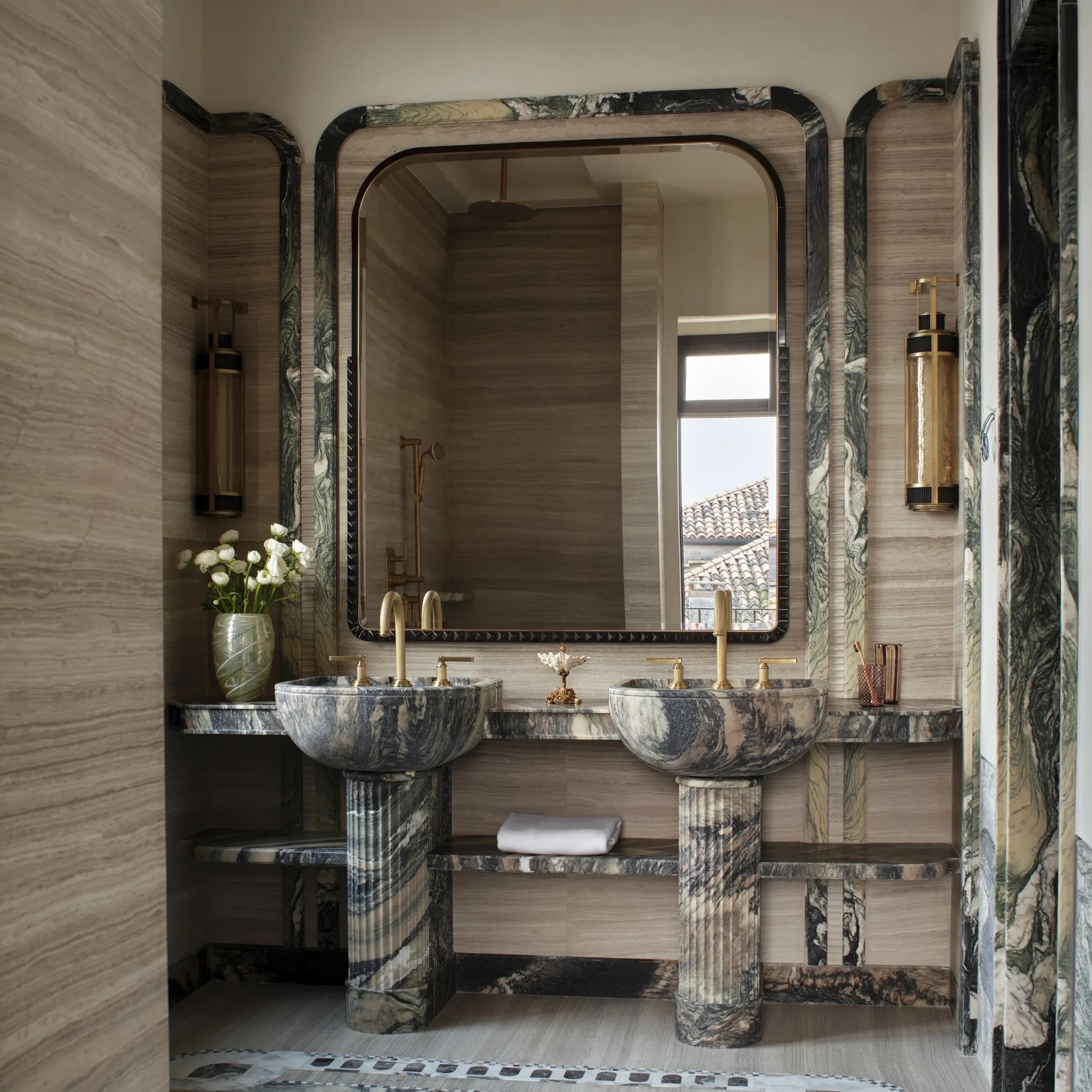A double marble vanity with veined green and black stone, brass fittings, round mirrors, and a vase of white ranunculus between the basins