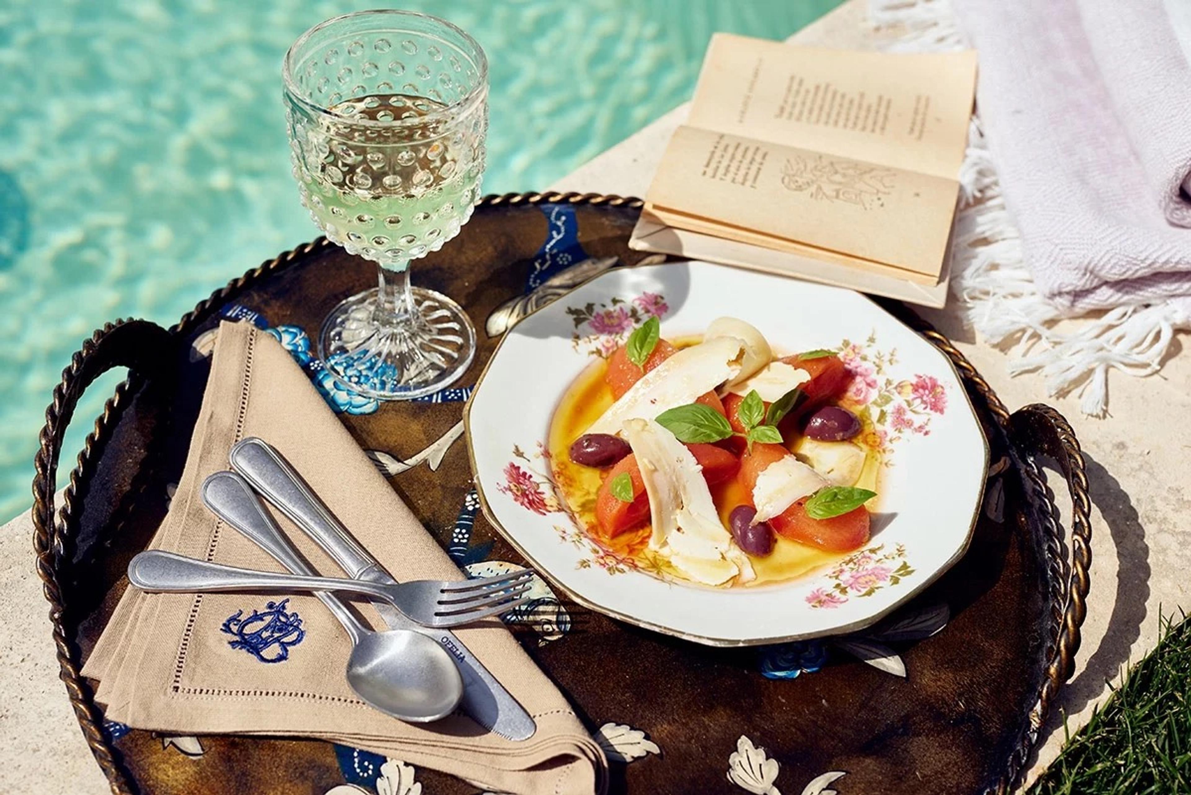 A plate of sliced tomato, soft cheese and olives on a tray beside a pool, with a glass of wine and an open book.