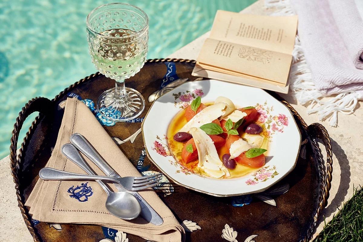 A plate of sliced tomato, soft cheese and olives on a tray beside a pool, with a glass of wine and an open book.