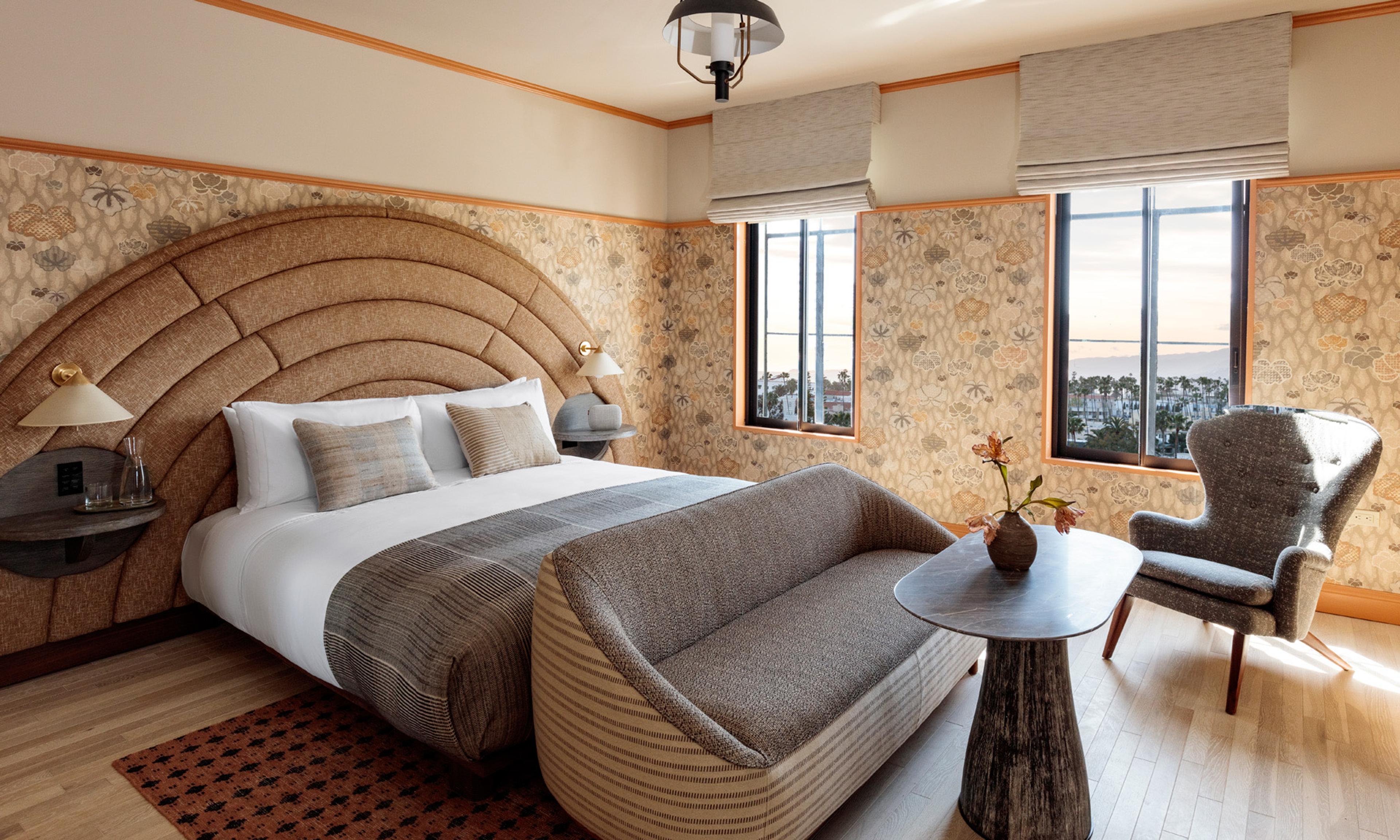 A hotel bedroom with a curved woven headboard, a grey striped bedcover, a sofa and armchair, and two windows looking out over the marina.