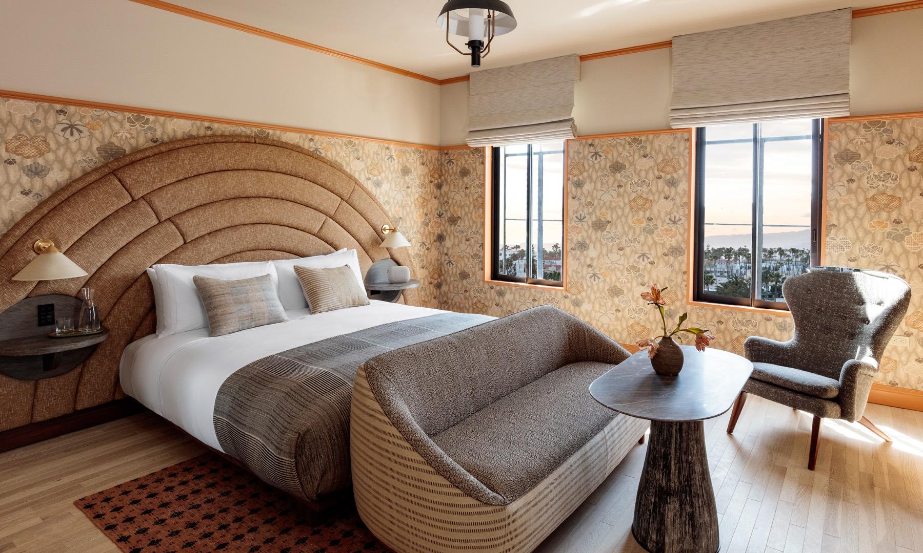 A hotel bedroom with a curved woven headboard, a grey striped bedcover, a sofa and armchair, and two windows looking out over the marina.